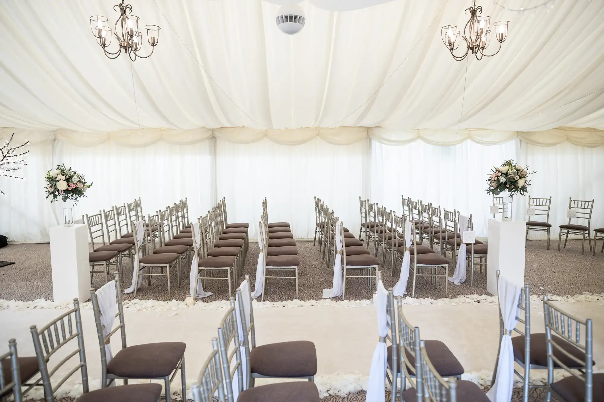 A wedding venue with rows of chairs facing an altar under a white tent, decorated with chandeliers and floral arrangements.