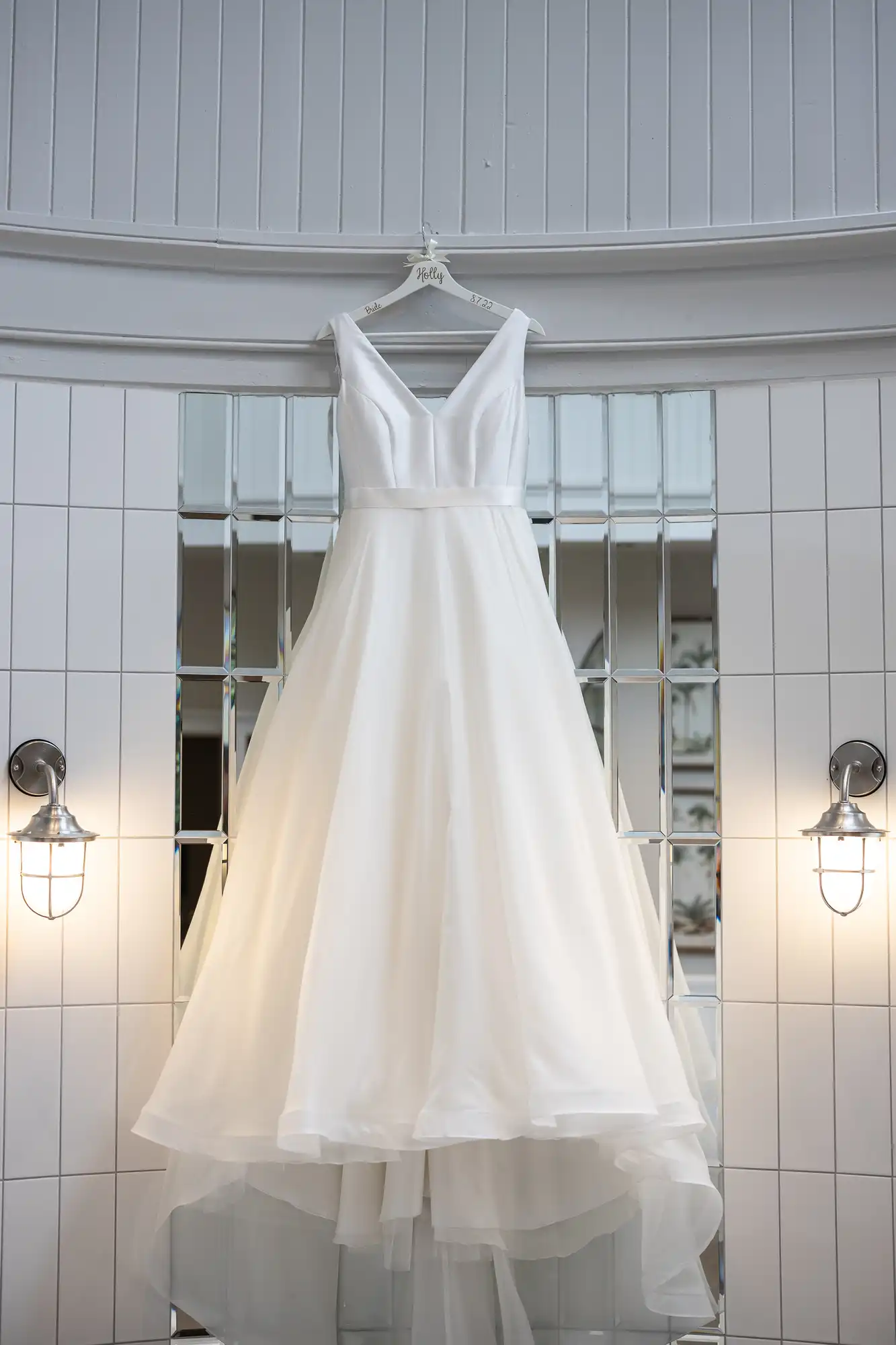 A white sleeveless wedding dress with a V-neckline and layered skirt is hanging on a white wooden hanger against a tiled wall with two sconces.