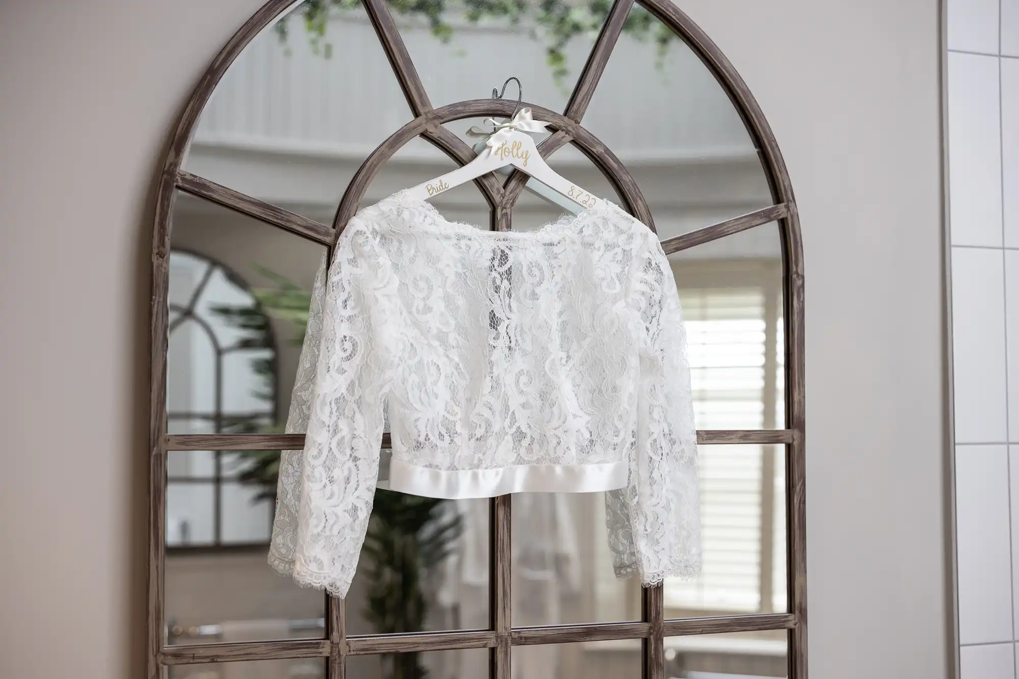 A white lace bridal top with a satin waistband and long sleeves is hung on a personalized hanger in front of a large, arched mirror.