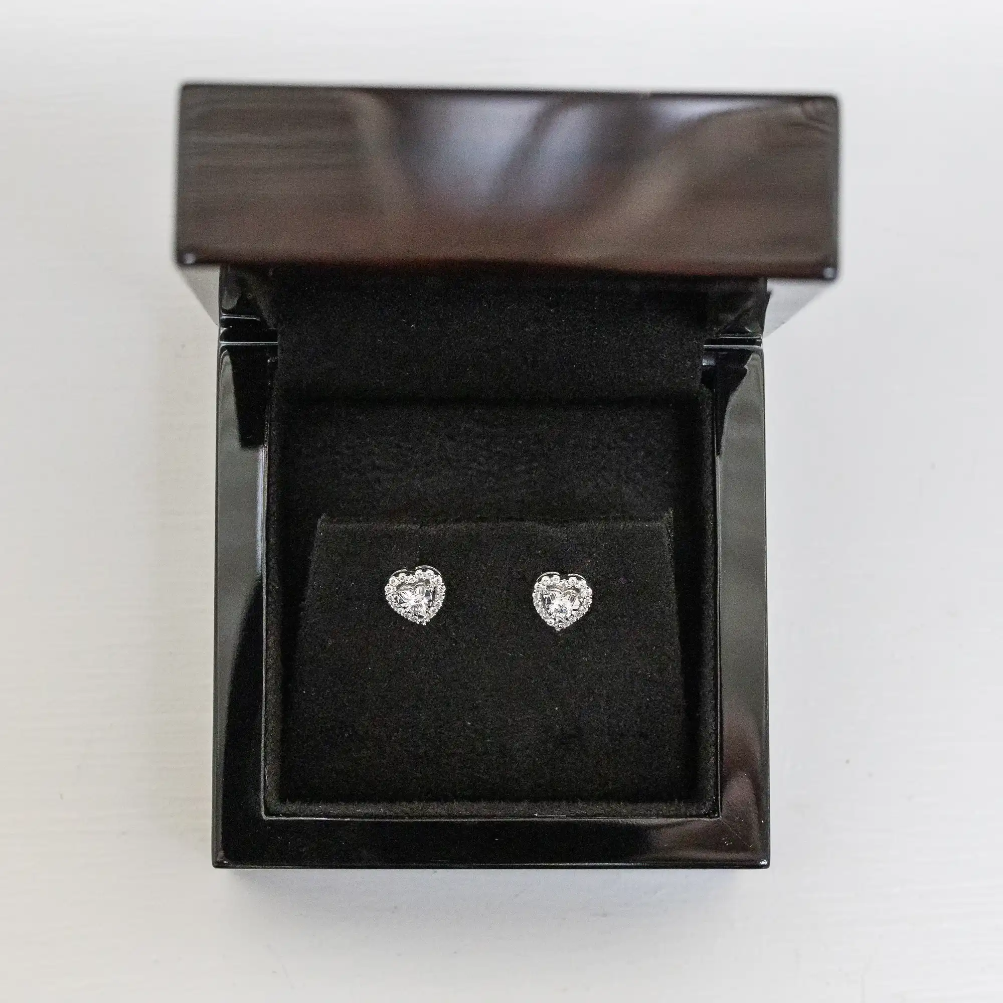 Pair of heart-shaped diamond earrings displayed in an open, black jewelry box with a velvet interior.