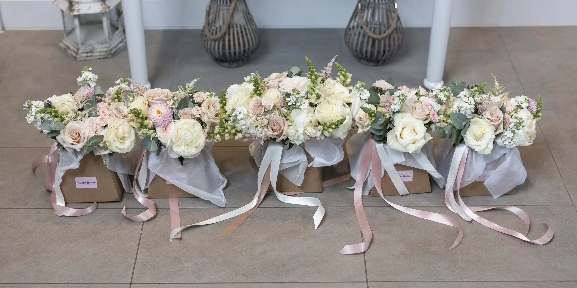 Several boxed flower arrangements with bouquets of white, pink, and cream flowers, each adorned with ribbons, are placed on a tiled floor.