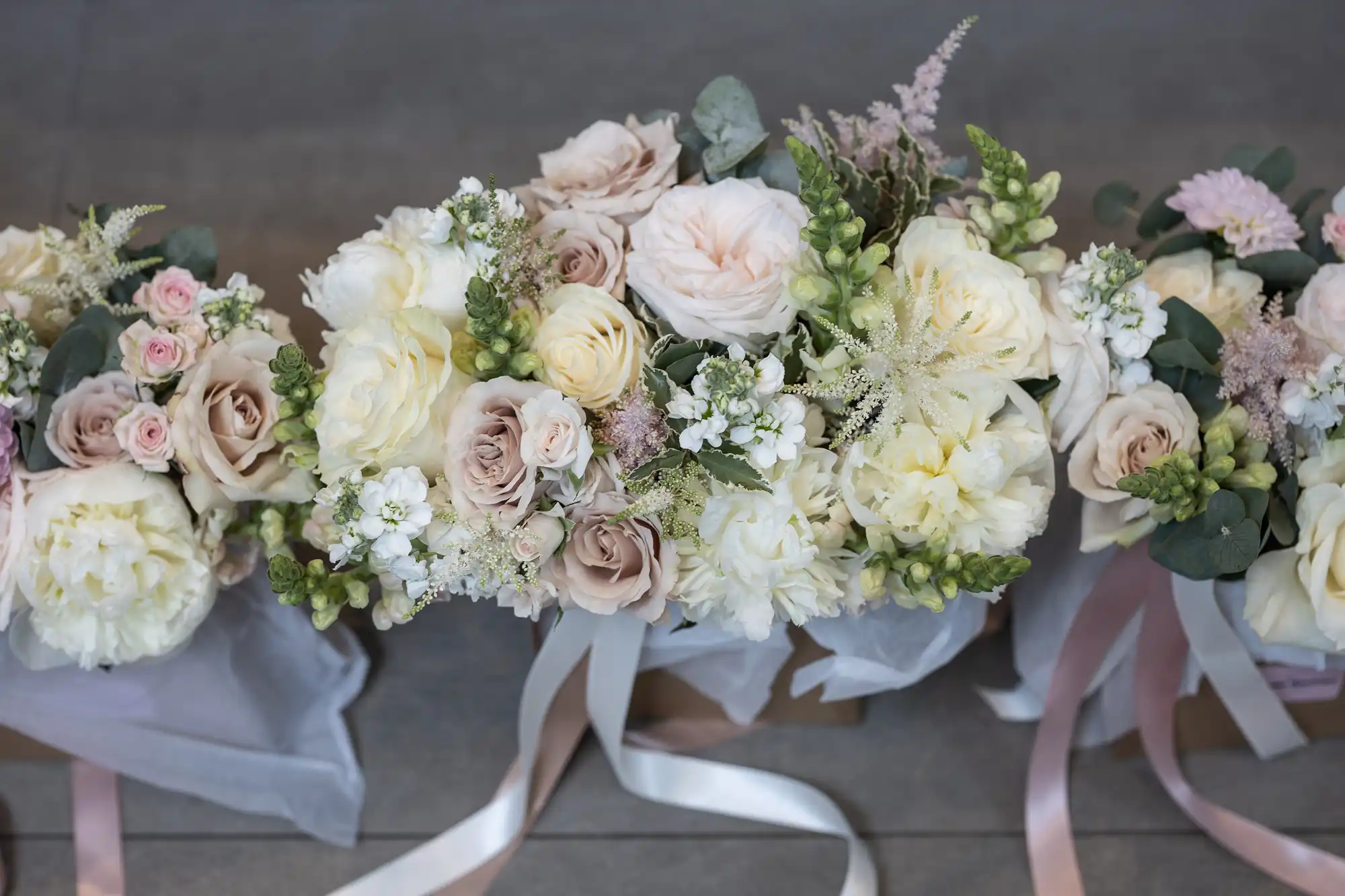 Bouquets of pastel-colored flowers, including roses and peonies, arranged with greenery and tied with white ribbons, placed on a gray surface.