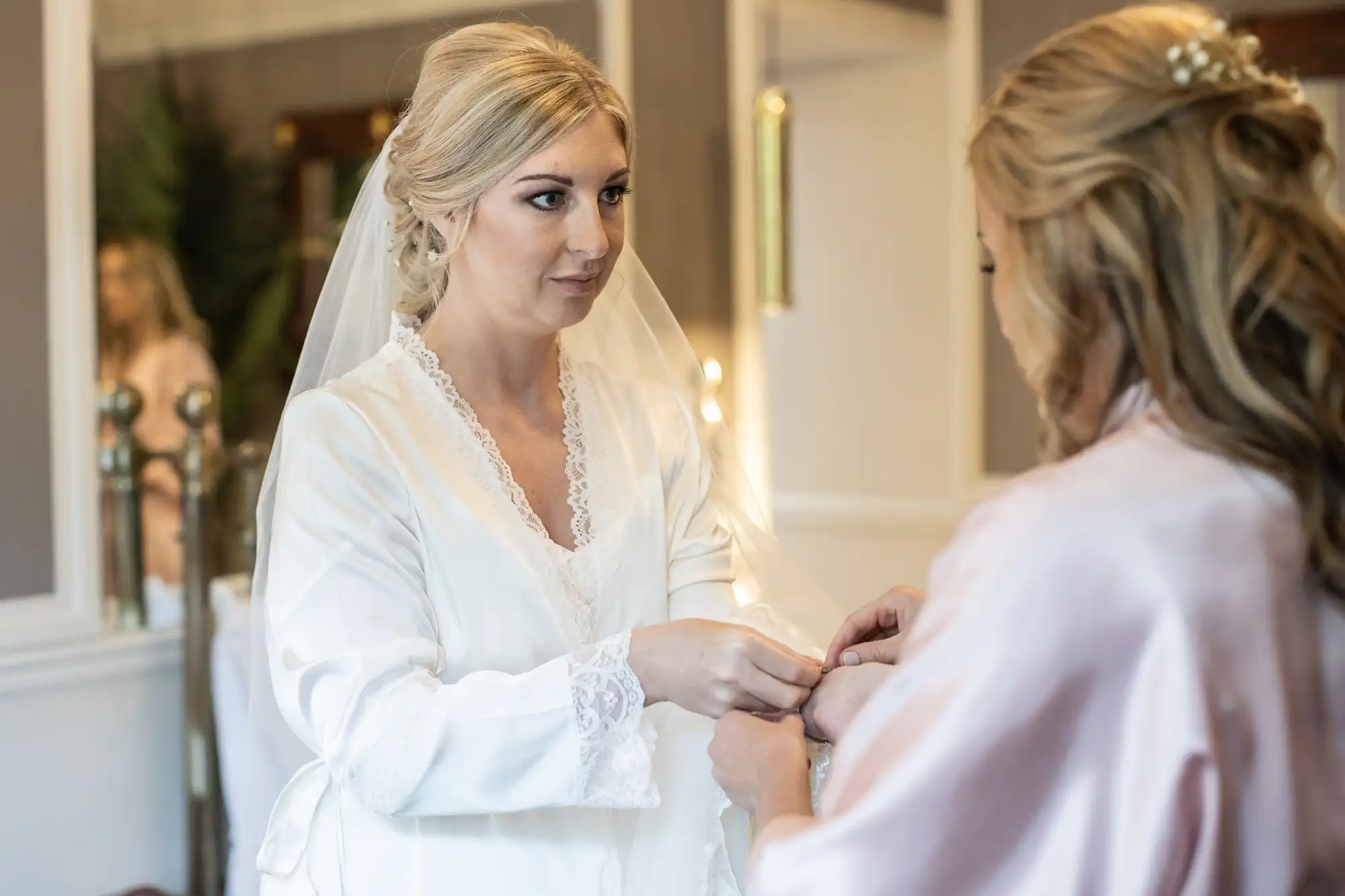 A bride in a white robe with lace trim holds hands with another woman with long hair, both standing indoors in a softly lit room.