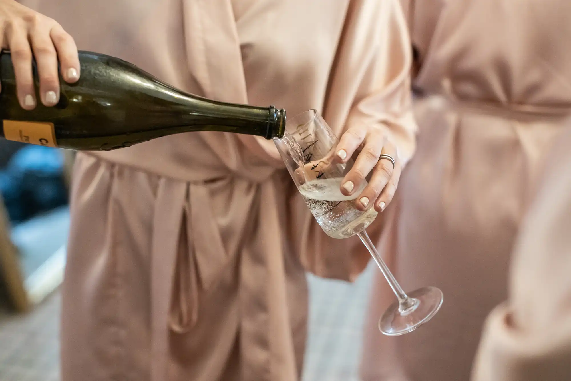 A person in a pink robe is pouring champagne into a glass held by another person in a similar robe.