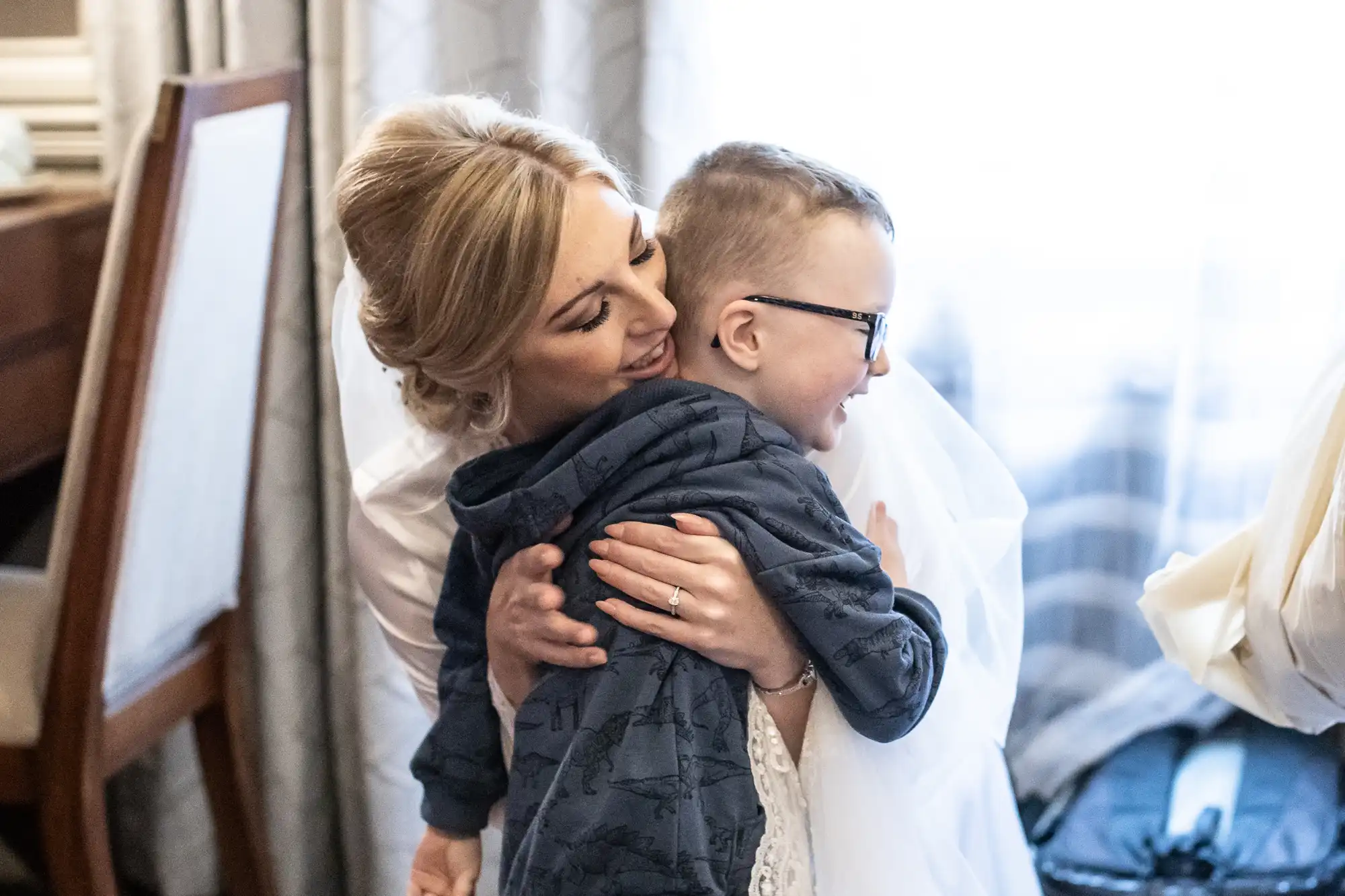 A woman with blonde hair hugs a child wearing glasses and a gray hoodie. They appear to share a moment of affection in what looks like a home setting.