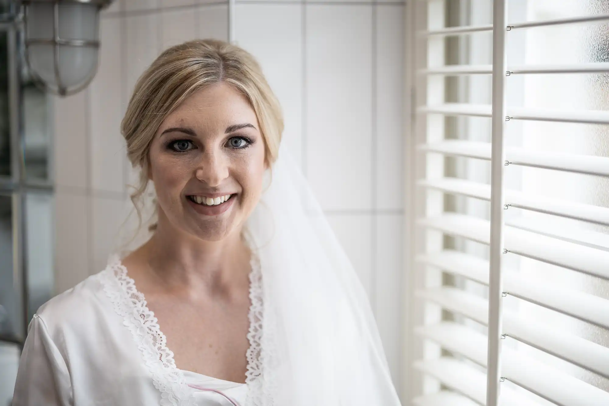 A woman with blonde hair wearing a white dress and veil smiles while standing indoors next to a window with blinds.