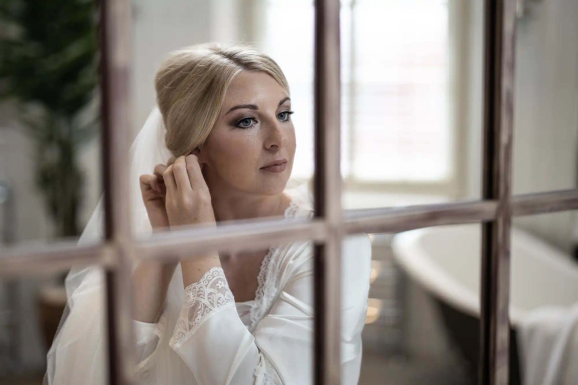 A bride wearing a white lace robe adjusts her earring while standing near a window.