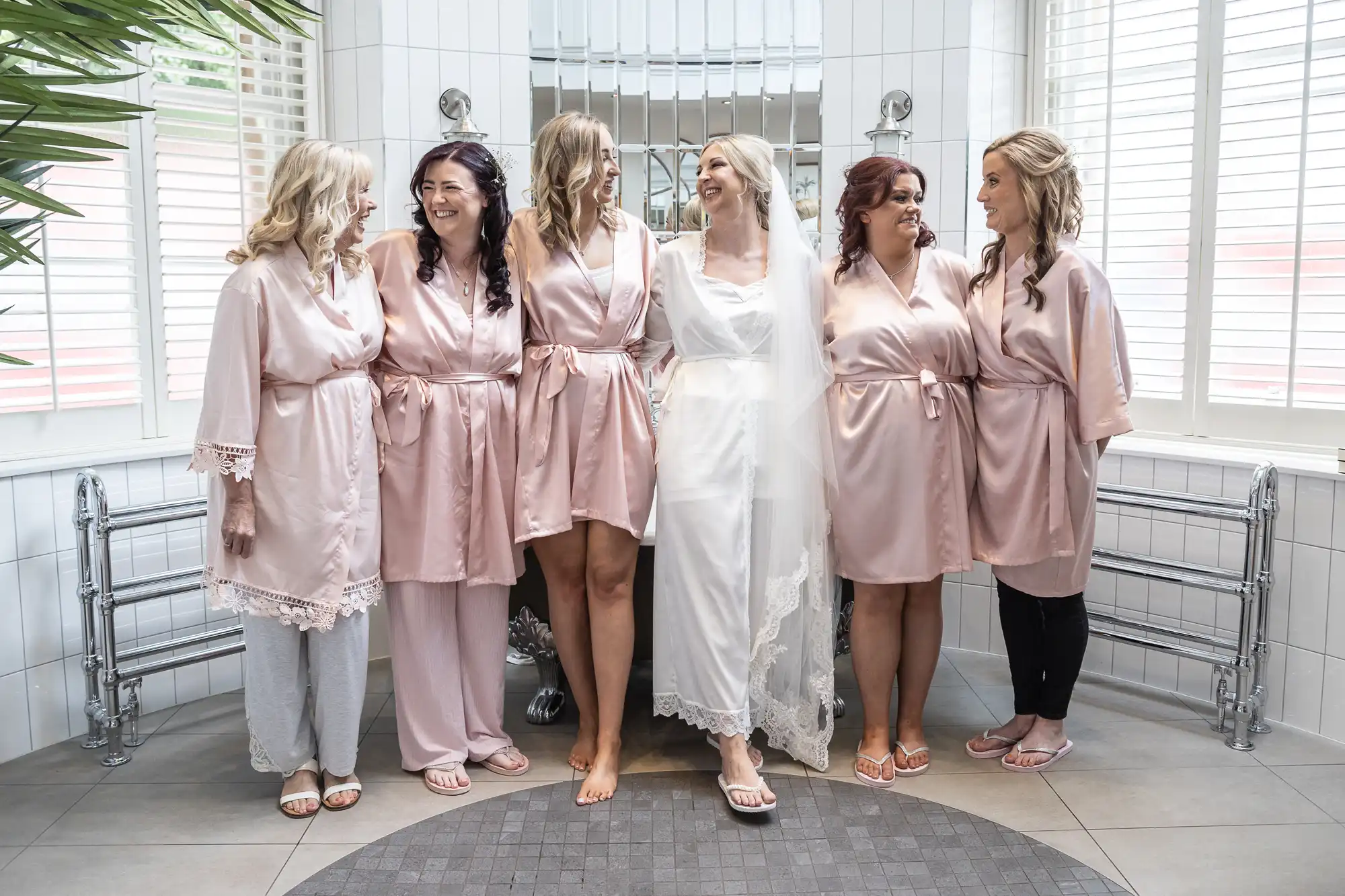 Six women in matching pink robes stand in a tiled room, smiling and looking at each other. The central woman is in a white robe with a veil.