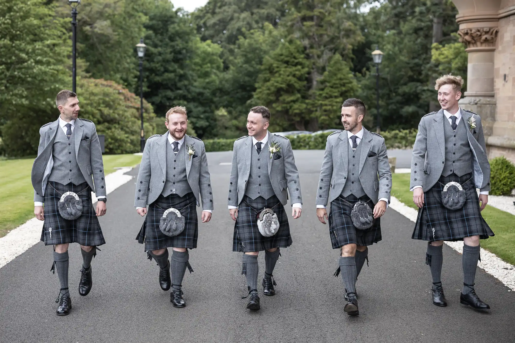 Five men in matching gray jackets, kilts, and sporrans walk on a paved path outside, surrounded by greenery. They are smiling and engaged in conversation.