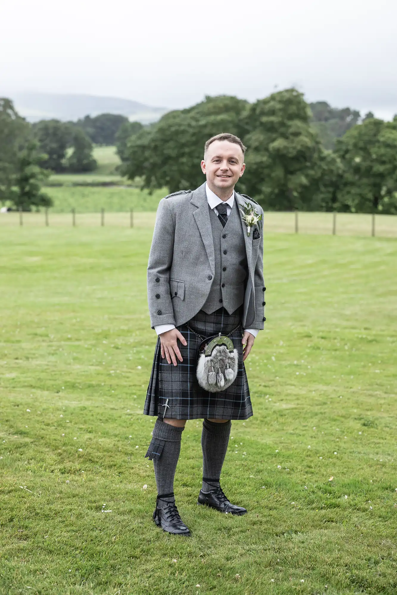 Man in traditional Scottish attire, including a grey jacket, a kilt, and knee-high socks, stands on a grassy field with trees and hills in the background.