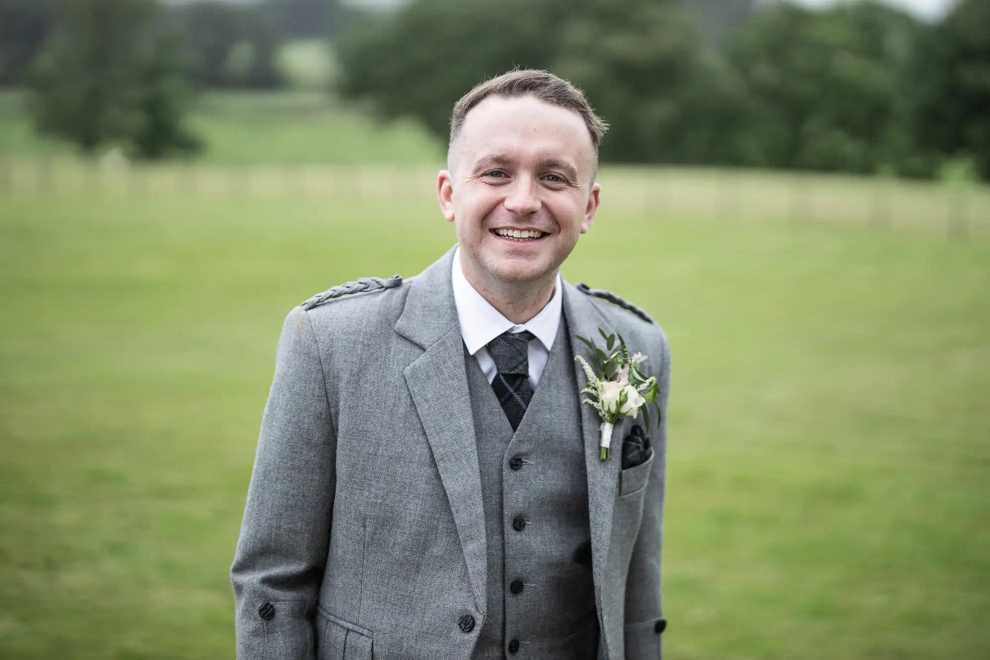 A man in a gray suit with a boutonniere stands outdoors on a grassy area, smiling at the camera.