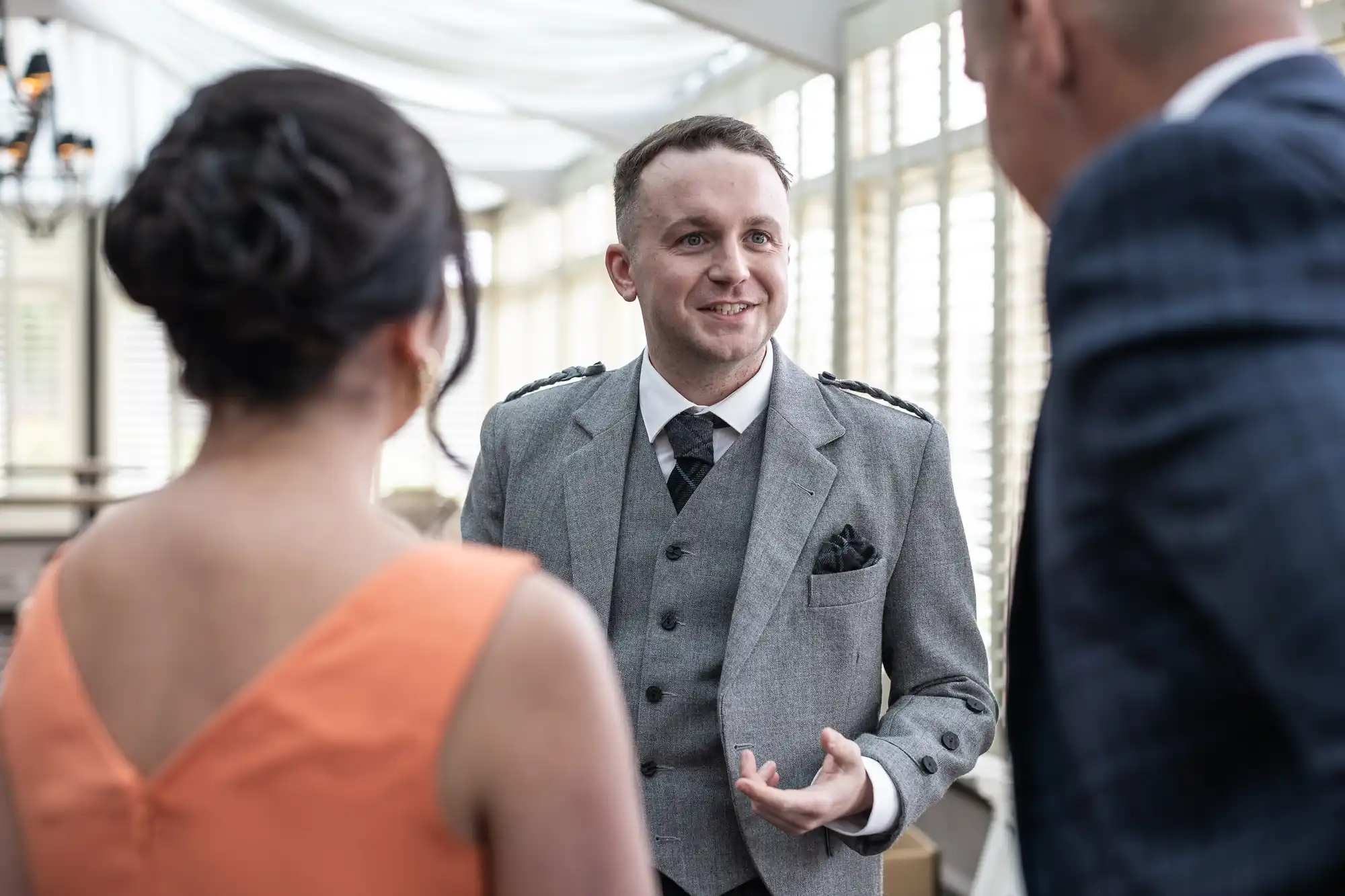 A man in a grey suit talks to a woman in an orange dress and another man in a dark suit inside a bright room.