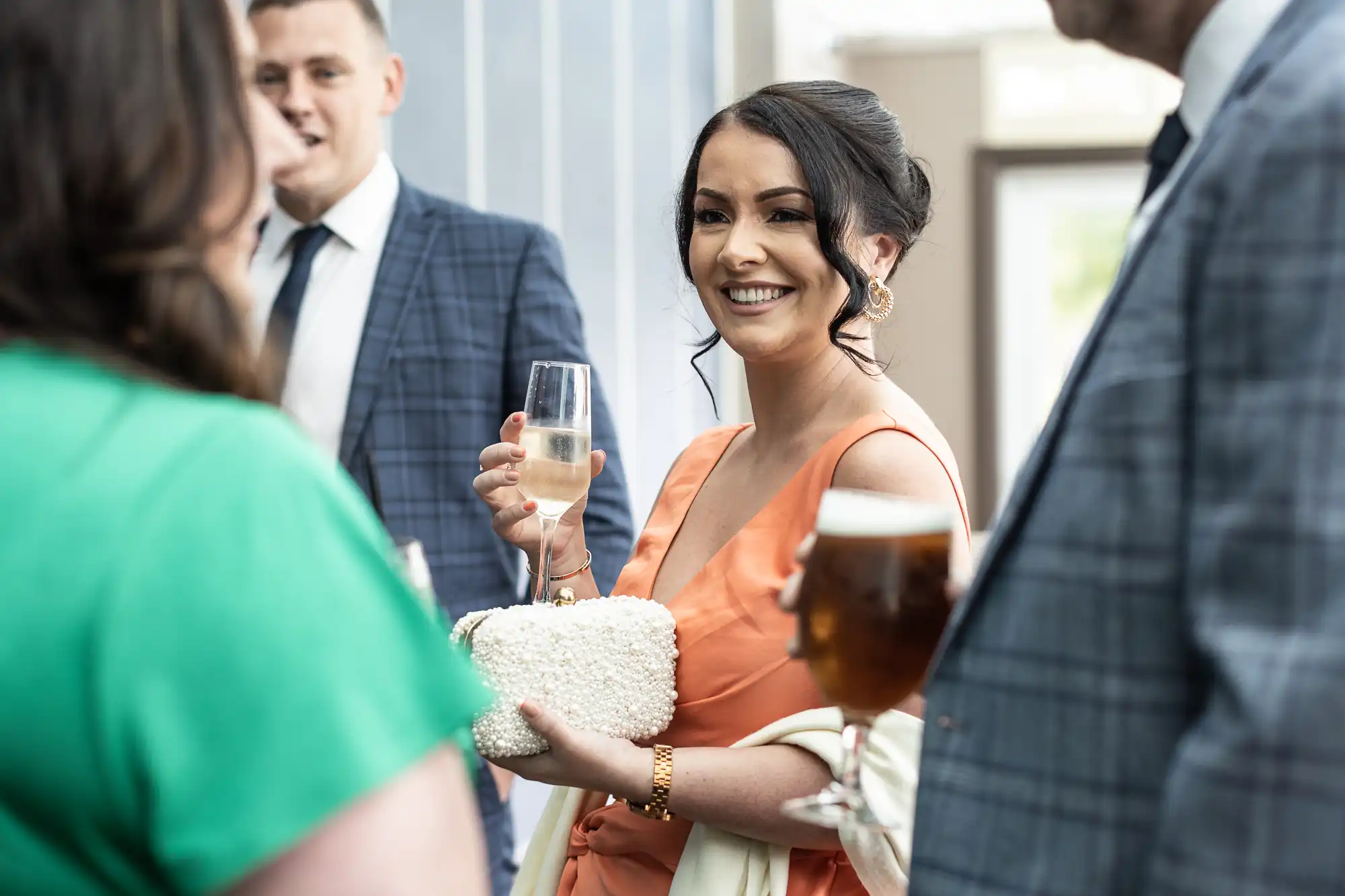 A woman in an orange dress holds a glass of champagne and a white clutch while conversing with people, including a man in a checked suit holding a beer.