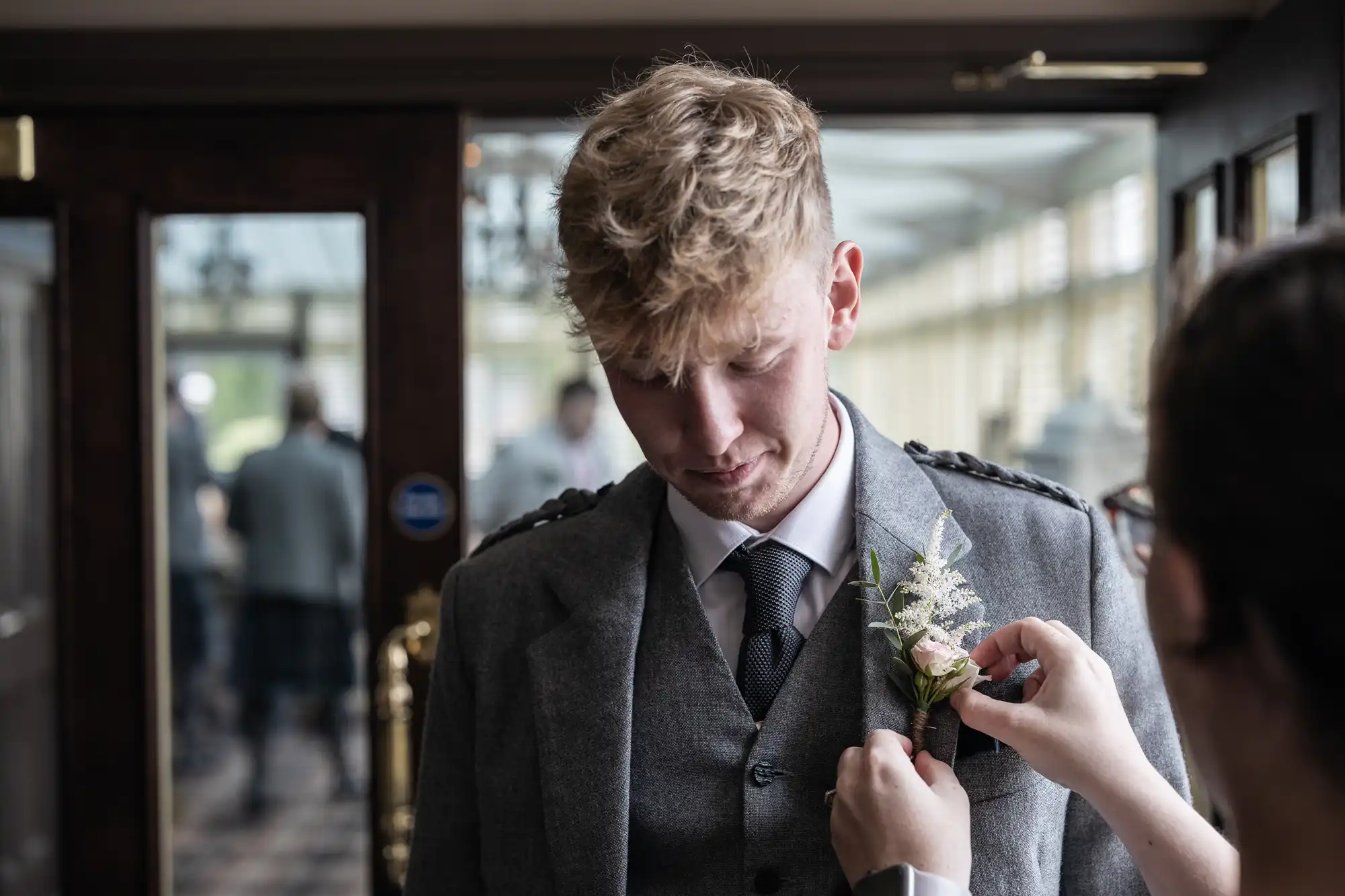 A person is adjusting a boutonniere on a man's suit jacket. The man is looking down, wearing a gray suit and a tie. They are indoors with blurred people and a door in the background.