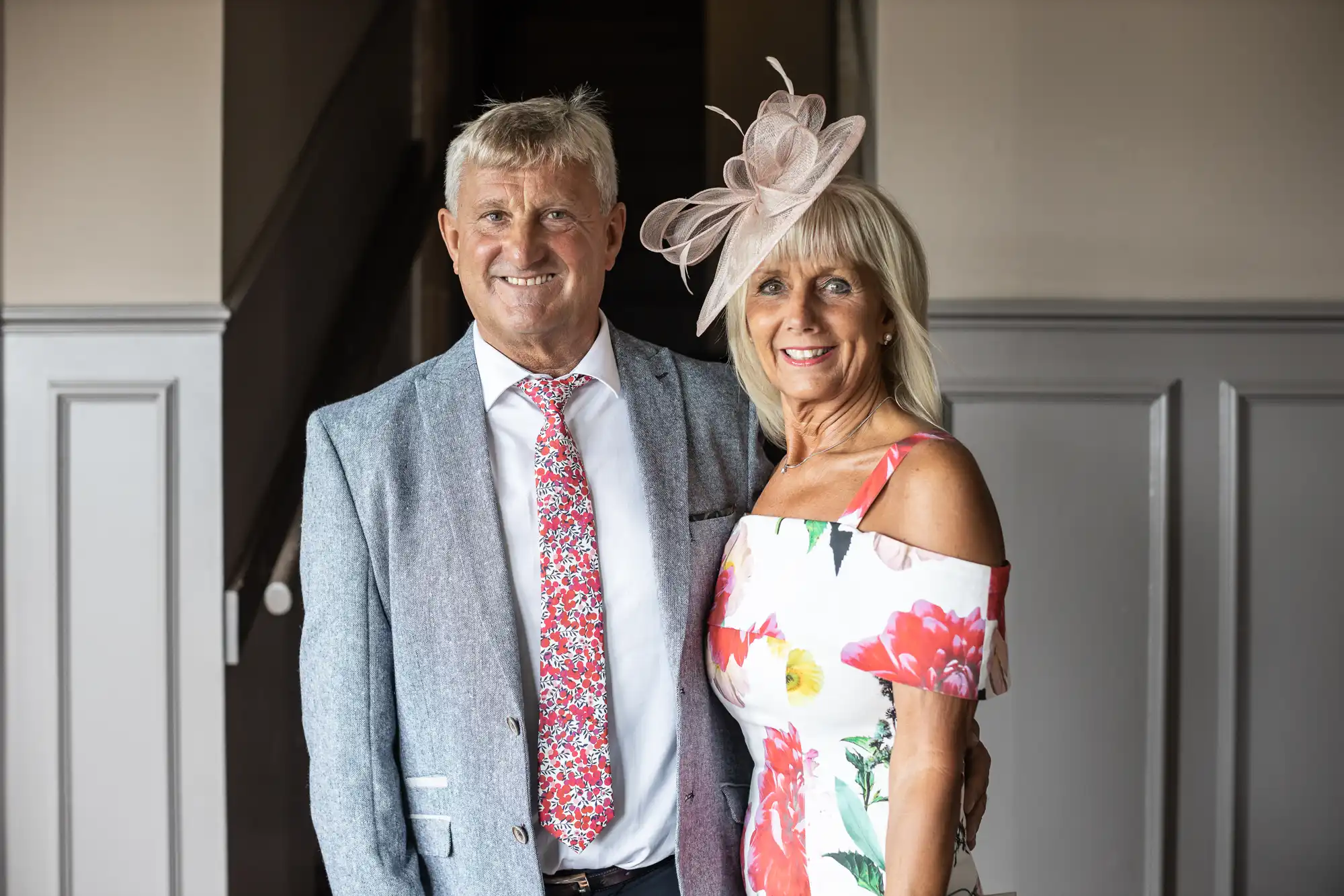 A man in a gray suit with a floral tie stands beside a woman in a floral dress and a hat, posing indoors.