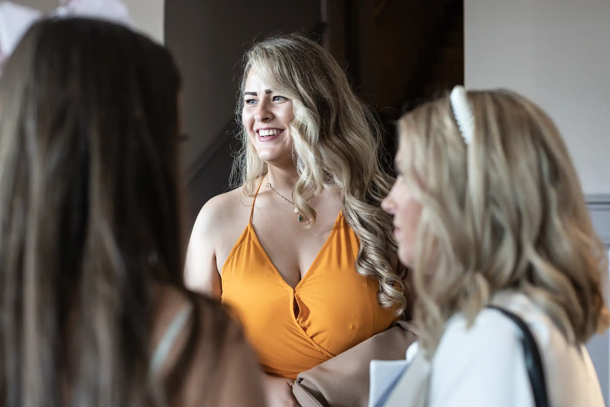 Three women are conversing indoors; the woman in an orange dress is smiling while the others are turned to the side.