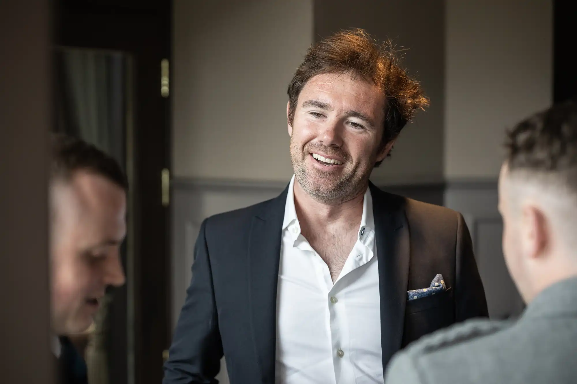 A man in a dark suit jacket and white shirt stands indoors, smiling and engaging in conversation with two other men.