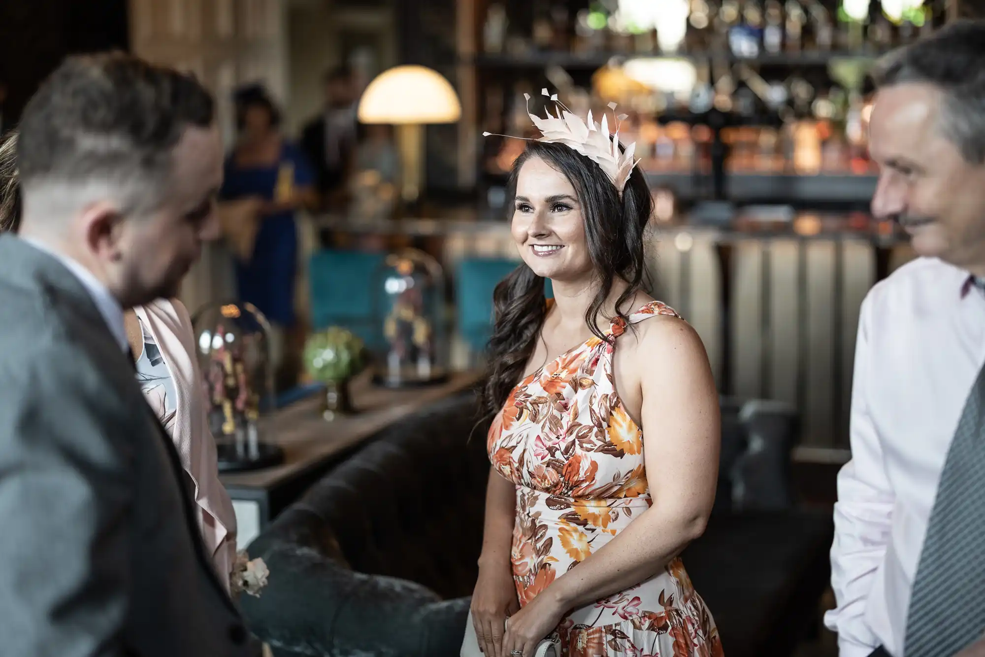 A woman in a floral dress and fascinator stands smiling among guests in a social setting.