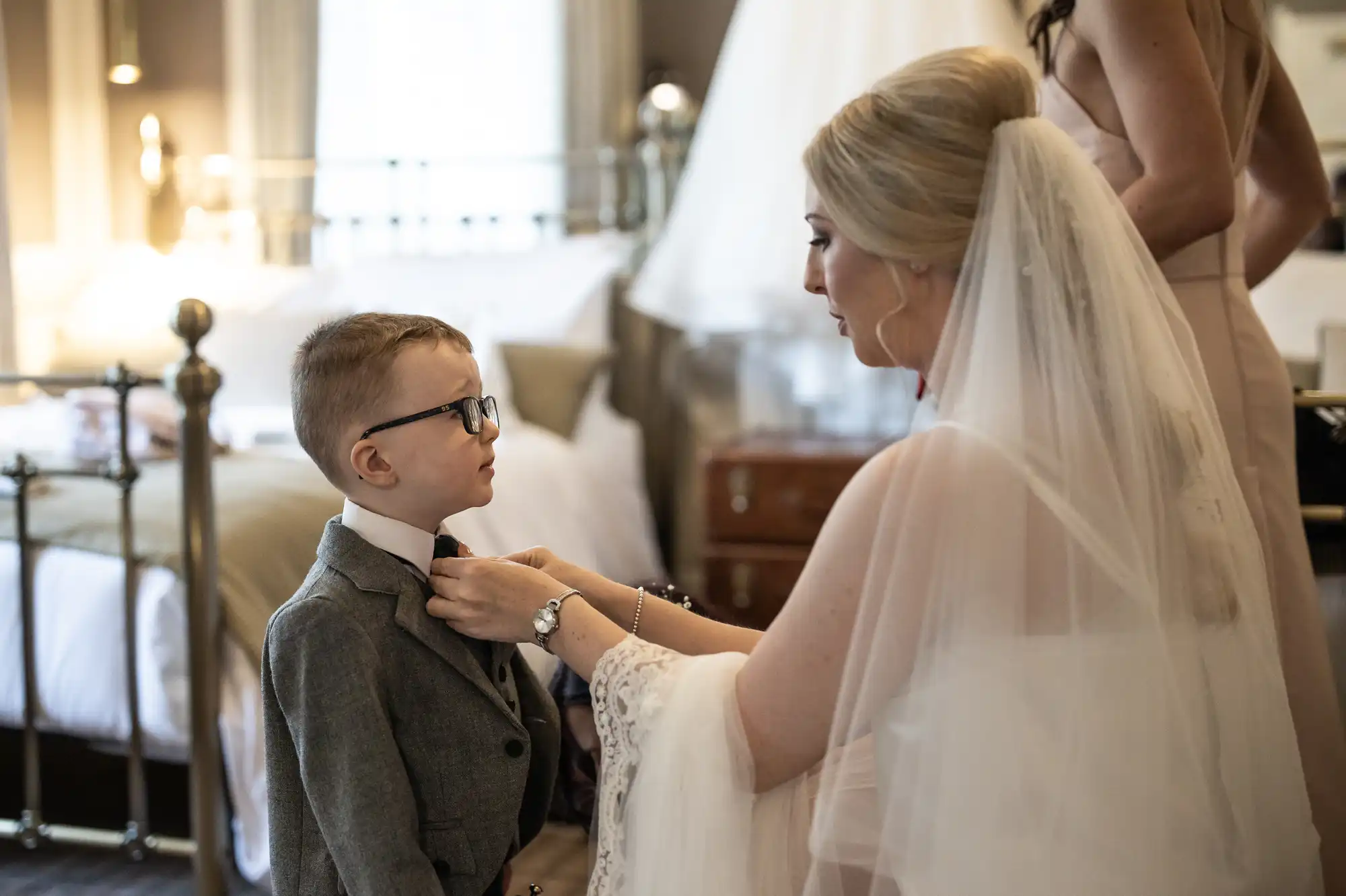 A bride is adjusting the bow tie of a young boy in a suit, standing in a bedroom with another person in the background.