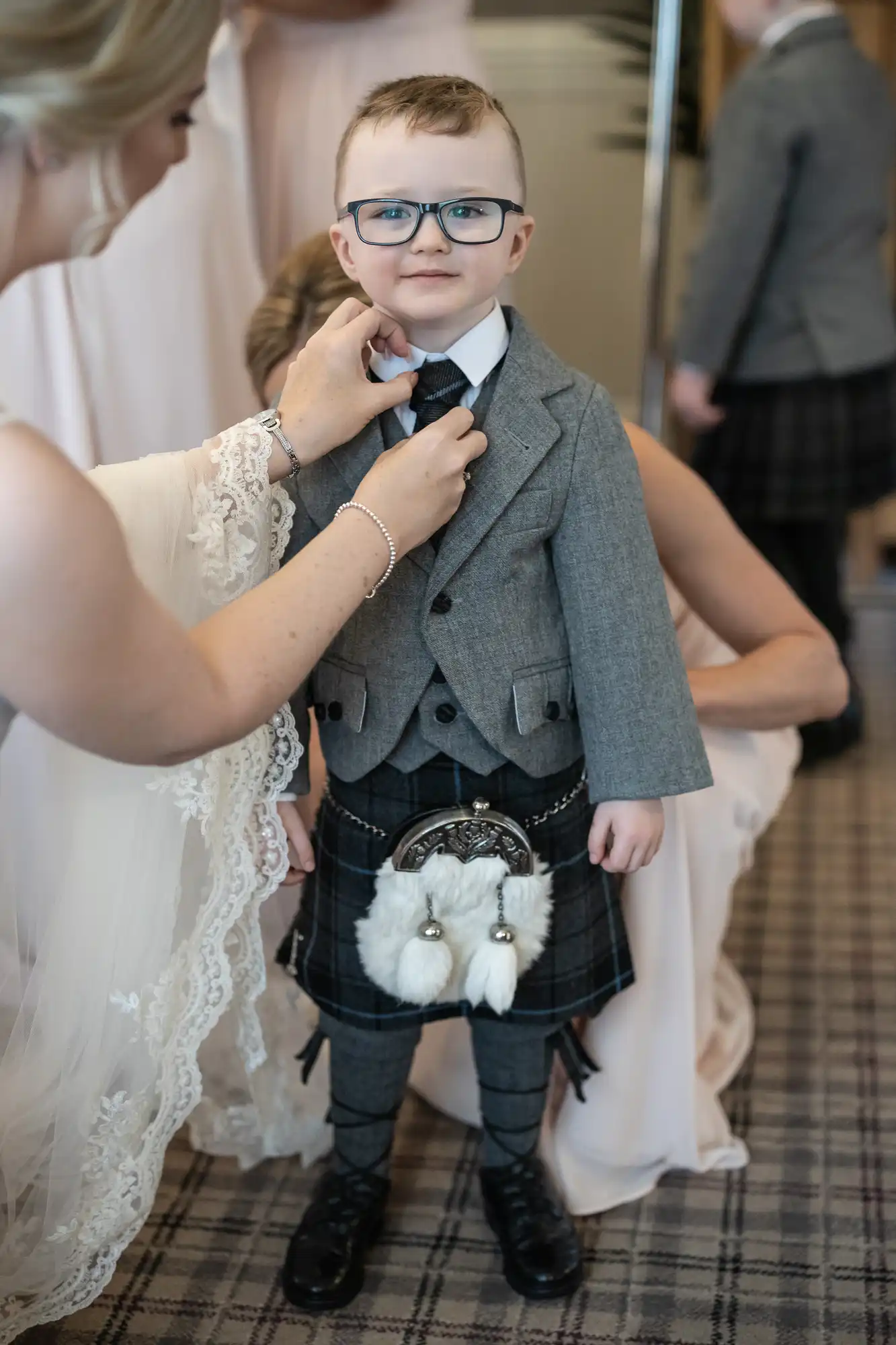 A young boy wearing glasses and traditional Scottish attire is having his tie adjusted by an adult. He is dressed in a grey jacket, white shirt, and kilt.