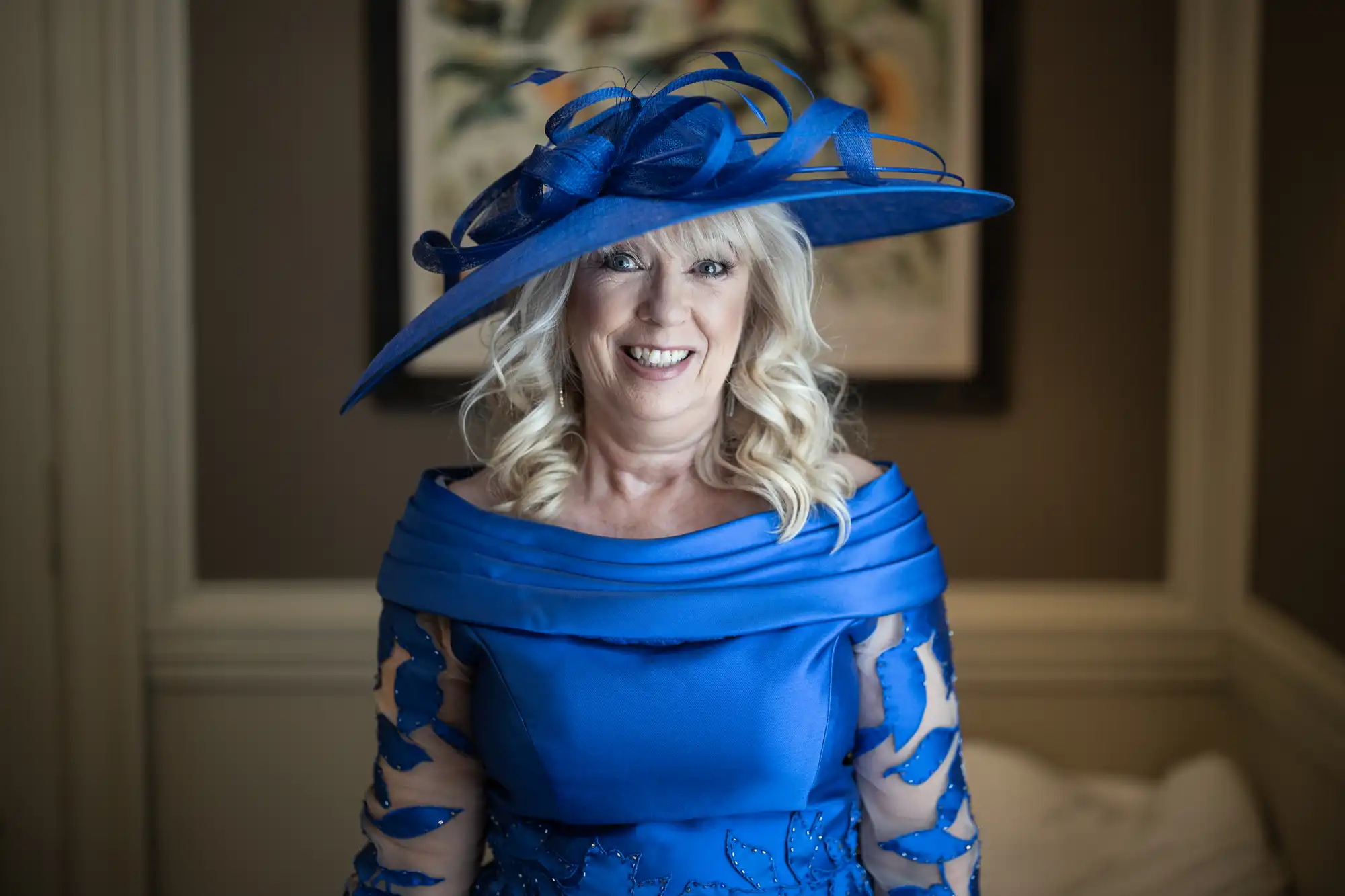 A woman with blonde hair wearing a blue dress and large matching blue hat stands indoors, smiling at the camera.