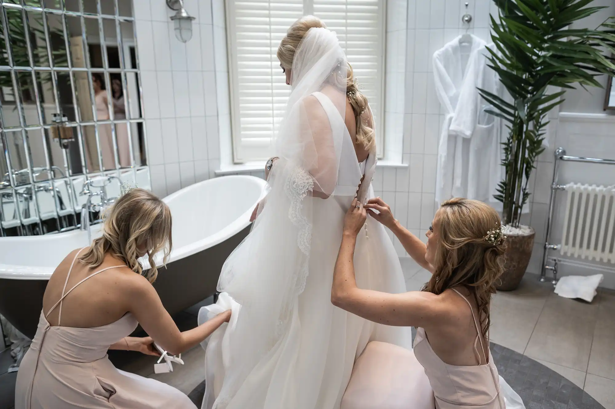 Two women assist a bride in adjusting her wedding dress in a bathroom. The bride is standing near a bathtub, and there is a large mirror and plant in the background.