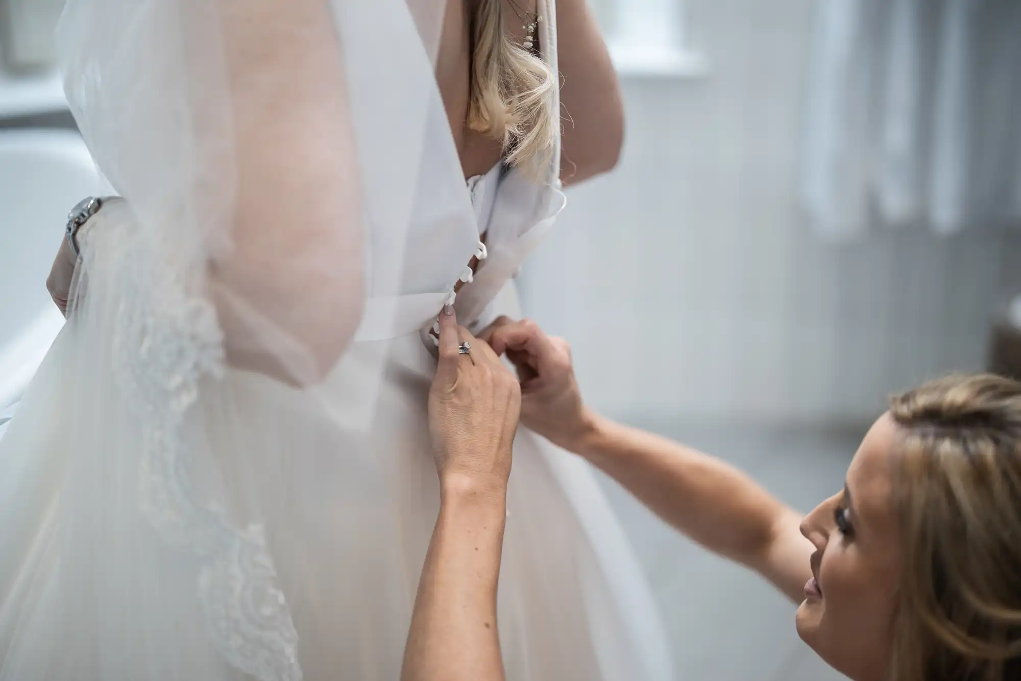 A person helps another with buttoning up a white wedding dress in a dimly lit room.