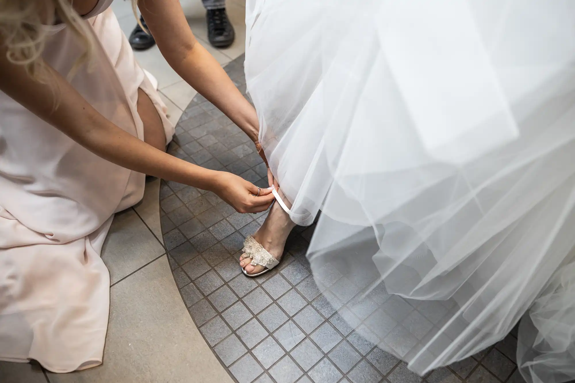 A person in a light pink dress adjusts the hem of a bride's white wedding gown while she wears white high-heeled shoes.