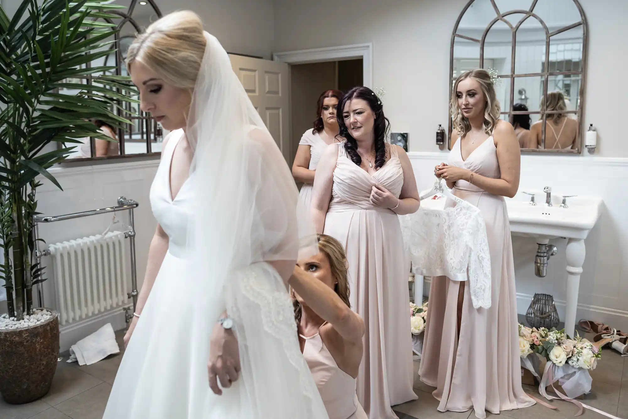 A bride in a white dress and veil prepares for her wedding day surrounded by bridesmaids in a room with large mirrors and a sink. One bridesmaid adjusts her dress, while another holds a lace garment.