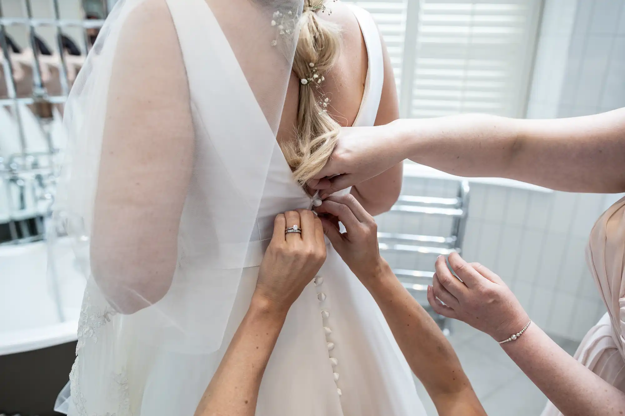 Two people help button up the back of a bride's white wedding dress. The scene is set indoors, with the bride's veil and part of a bathroom visible.