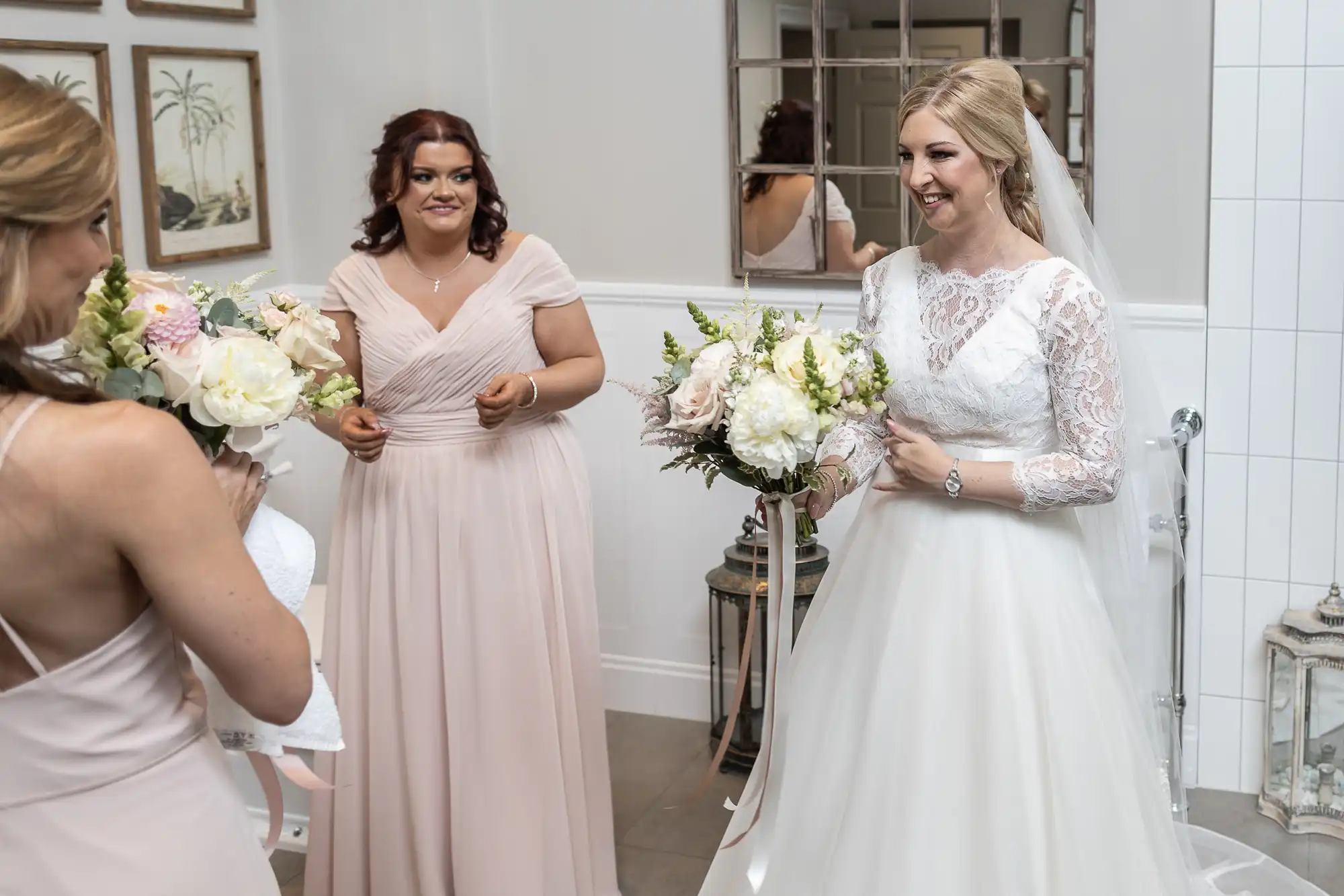 A bride in a white dress and veil holds a bouquet while smiling at two bridesmaids in light pink dresses who also hold bouquets. They are standing indoors, with framed pictures and a mirror in the background.