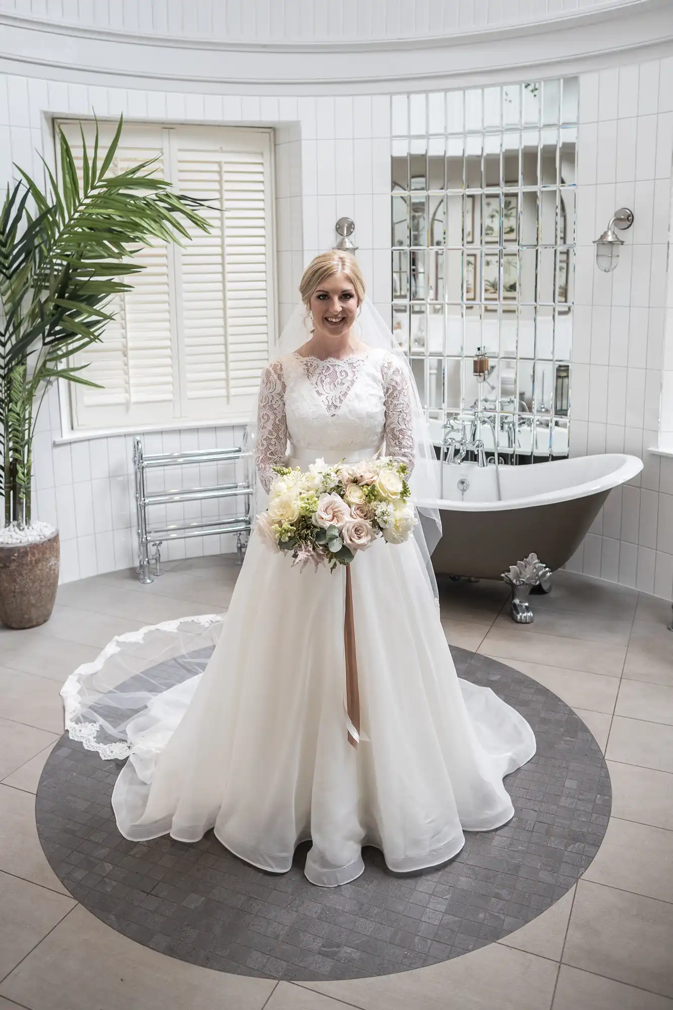 A bride in a lace wedding dress stands in front of a bathtub, holding a bouquet of flowers in a tiled bathroom with white walls and large windows.