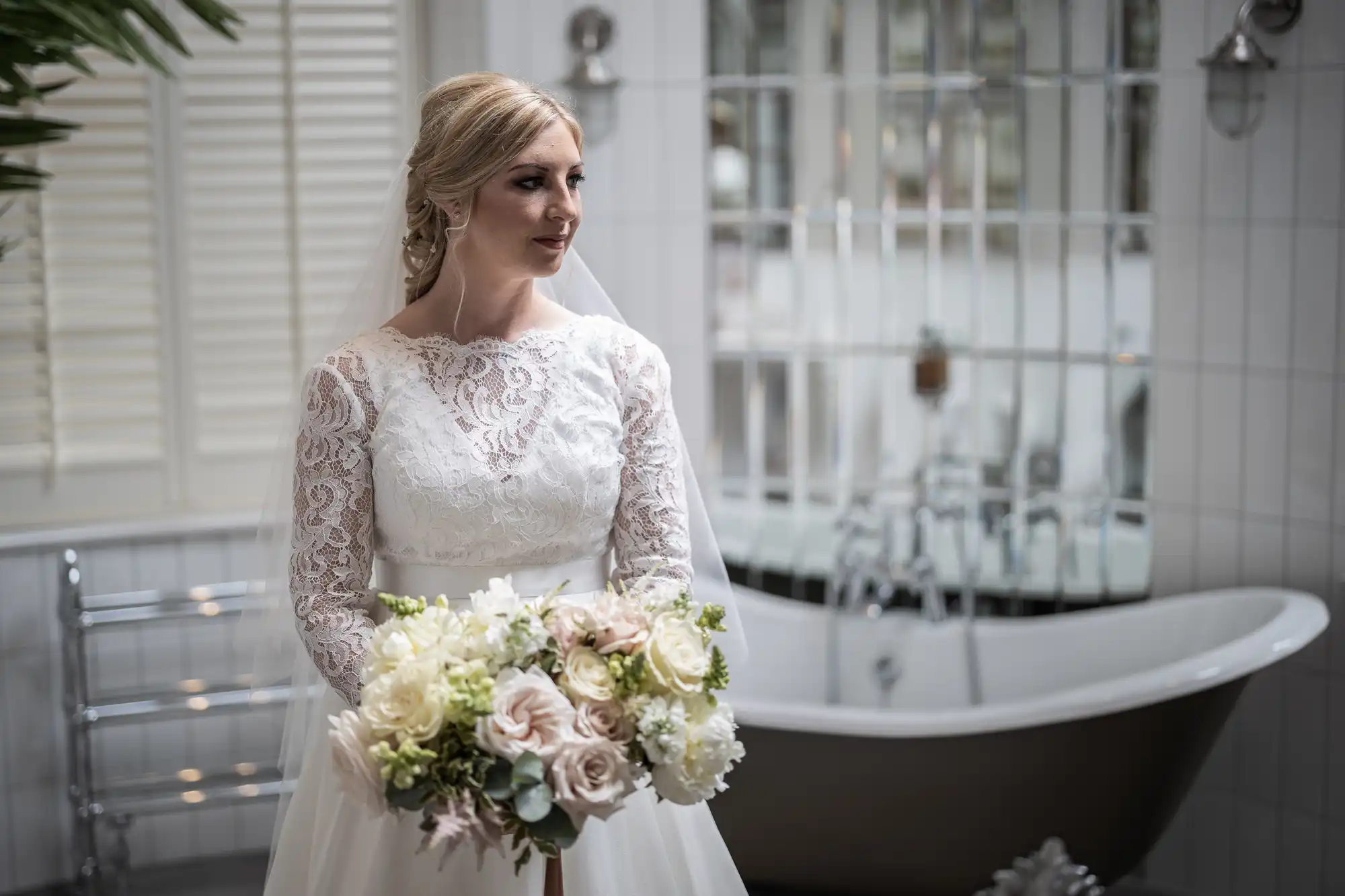 Bride in a white lace wedding dress holds a bouquet, standing in a bathroom with a clawfoot tub and tiled walls.