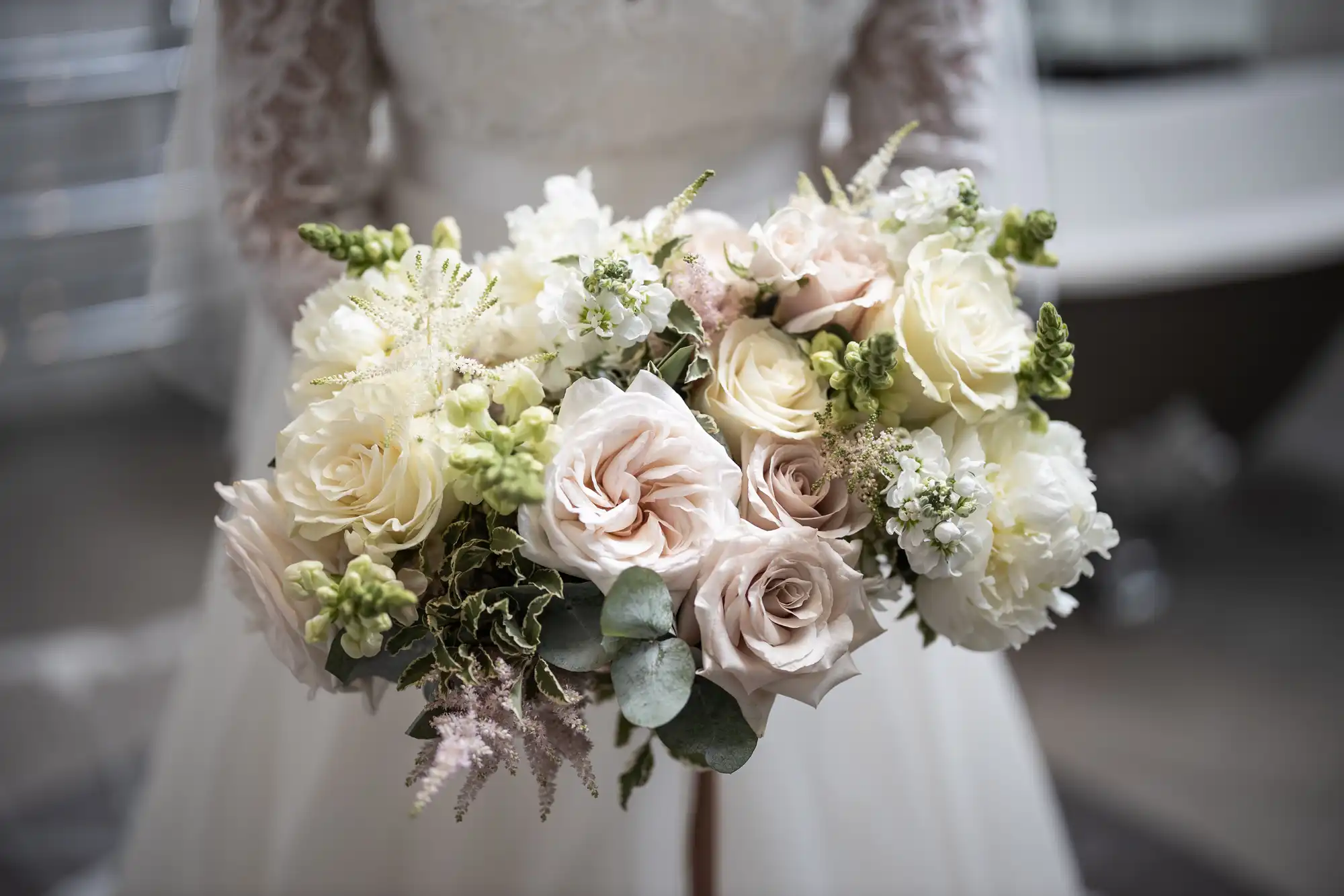 A bride holds a bouquet of white and pale pink flowers, including roses and greenery, while wearing a lace wedding dress.