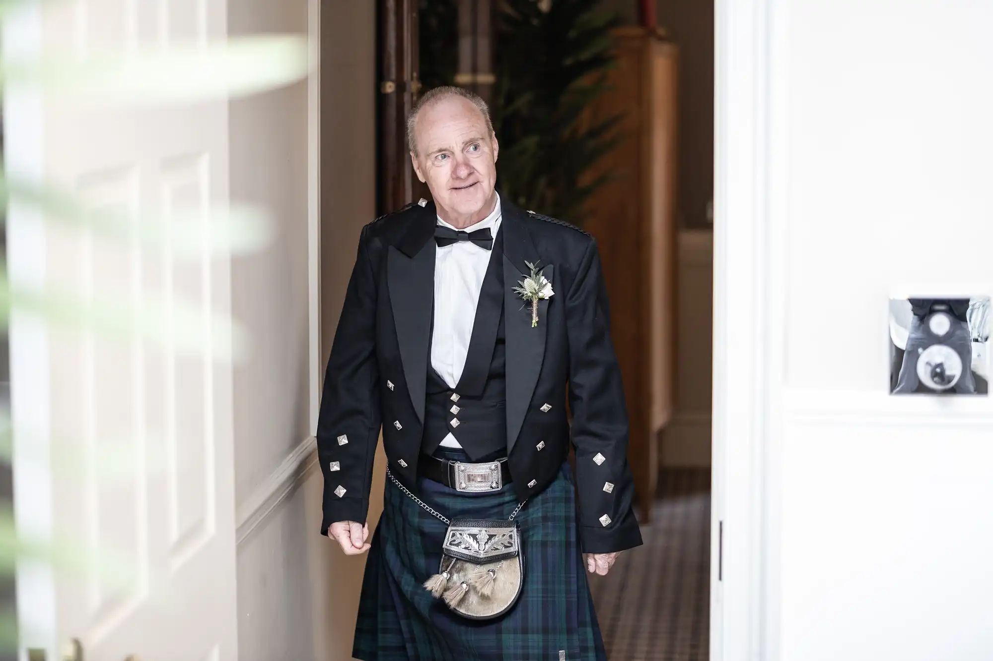 A man wearing a traditional Scottish kilt outfit stands in a doorway, smiling slightly, with a boutonniere on his suit jacket.