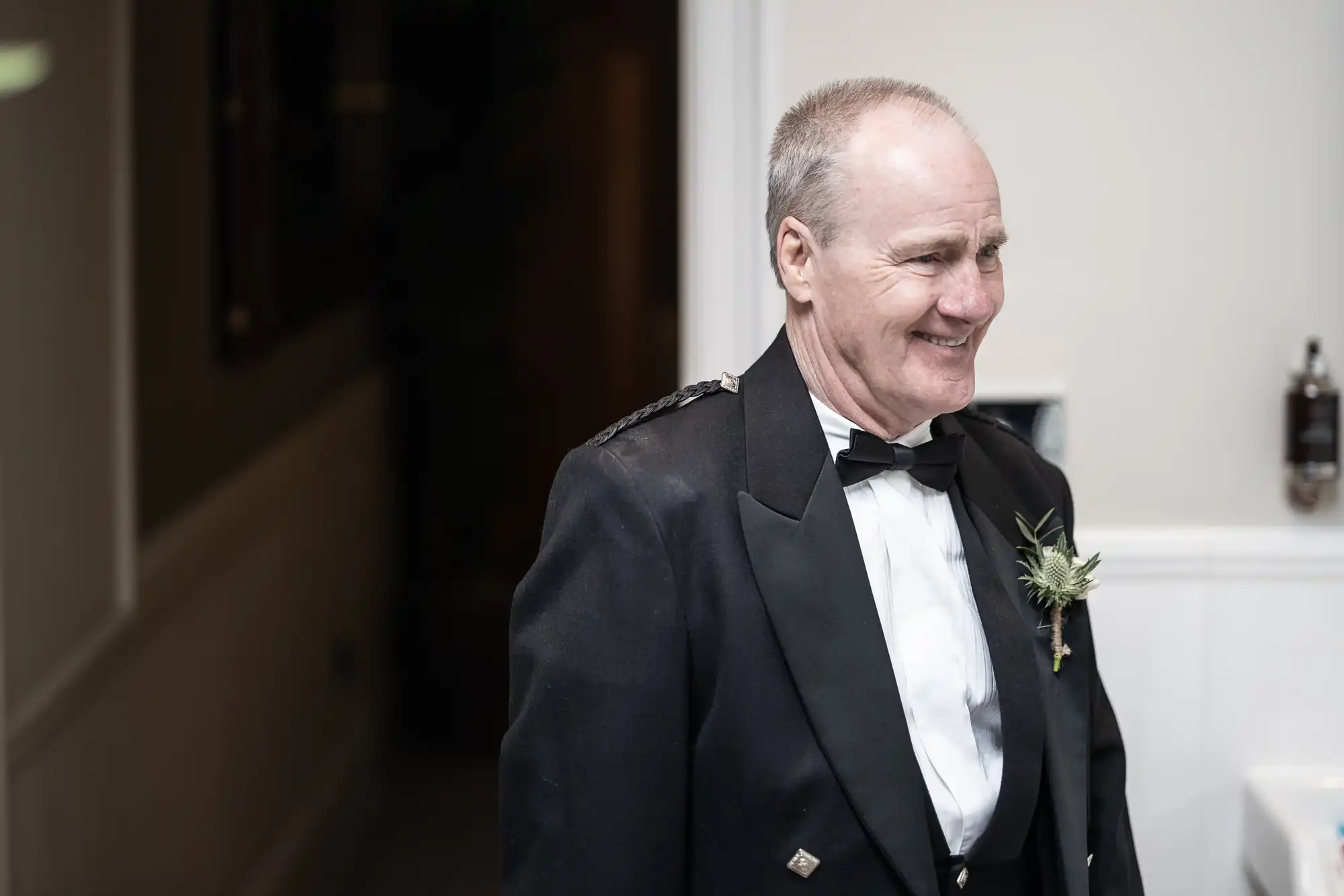 A man in formal attire, including a bow tie and kilt jacket, smiles while standing indoors. He has a boutonniere pinned to his jacket.