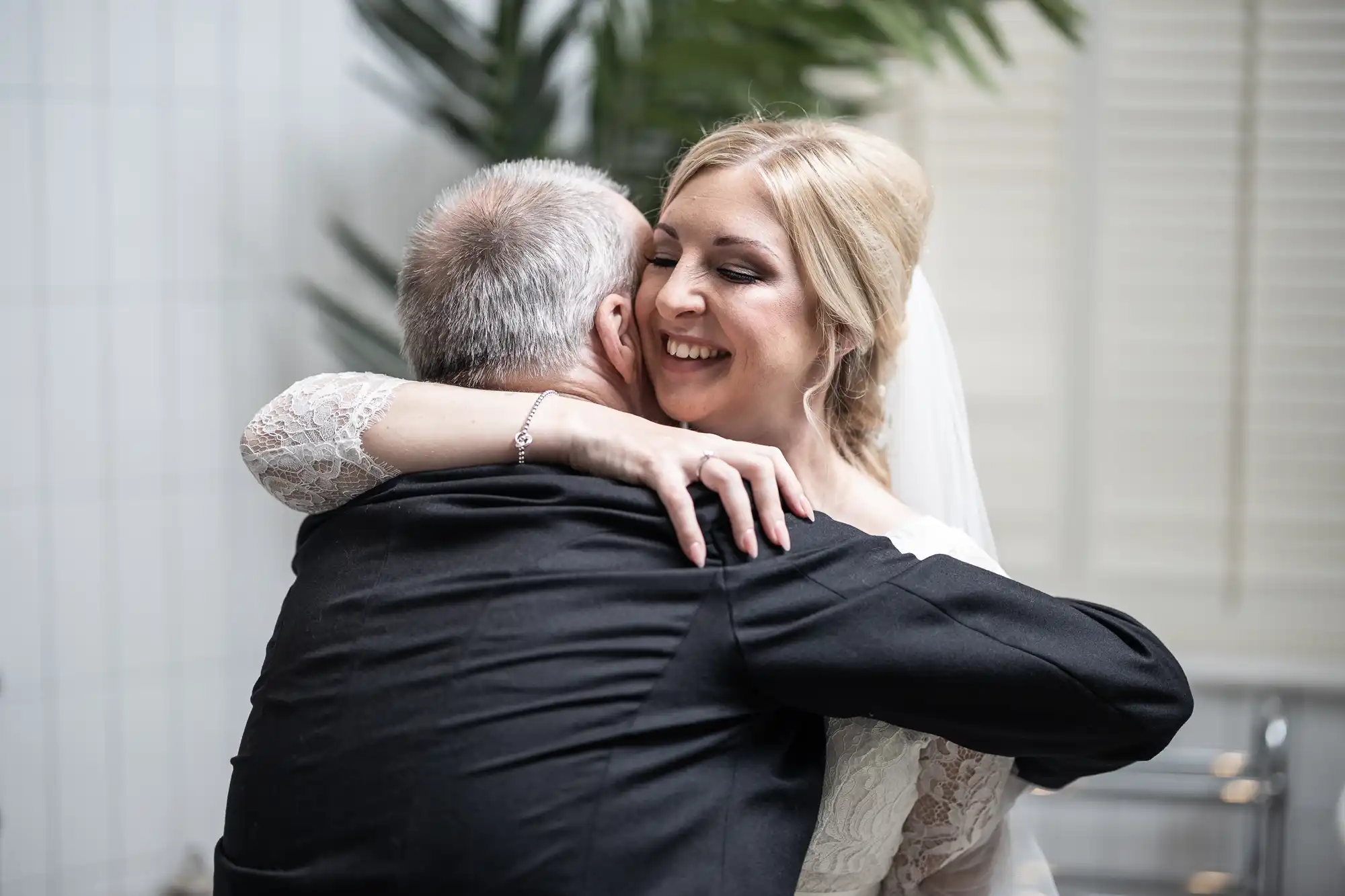 A bride in a white dress and veil hugs an older man in a suit, both smiling.