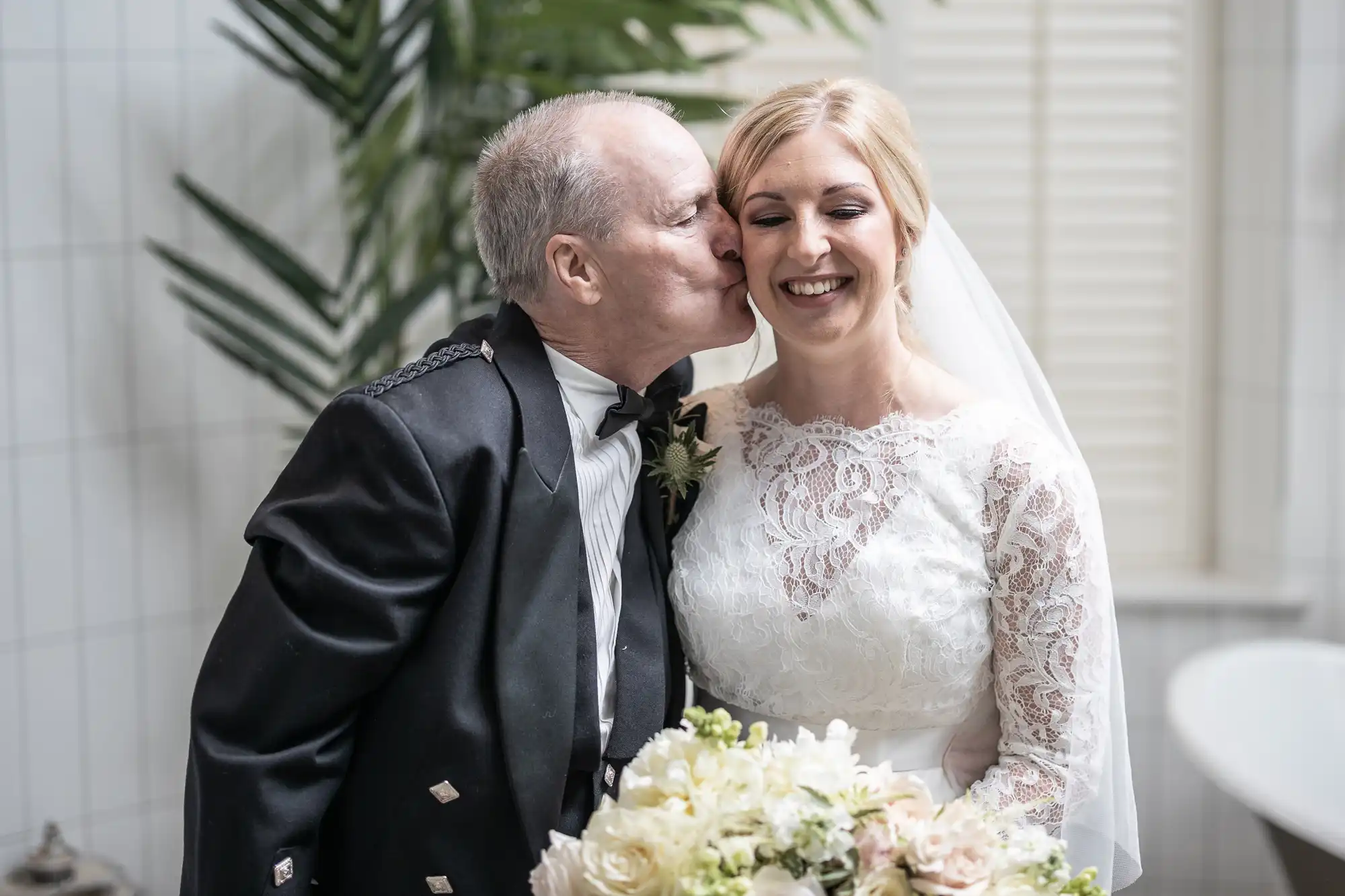 A man in formal attire kisses a smiling bride on her cheek. She is holding a bouquet and wearing a white lace wedding dress with a veil. They are standing indoors next to some greenery.