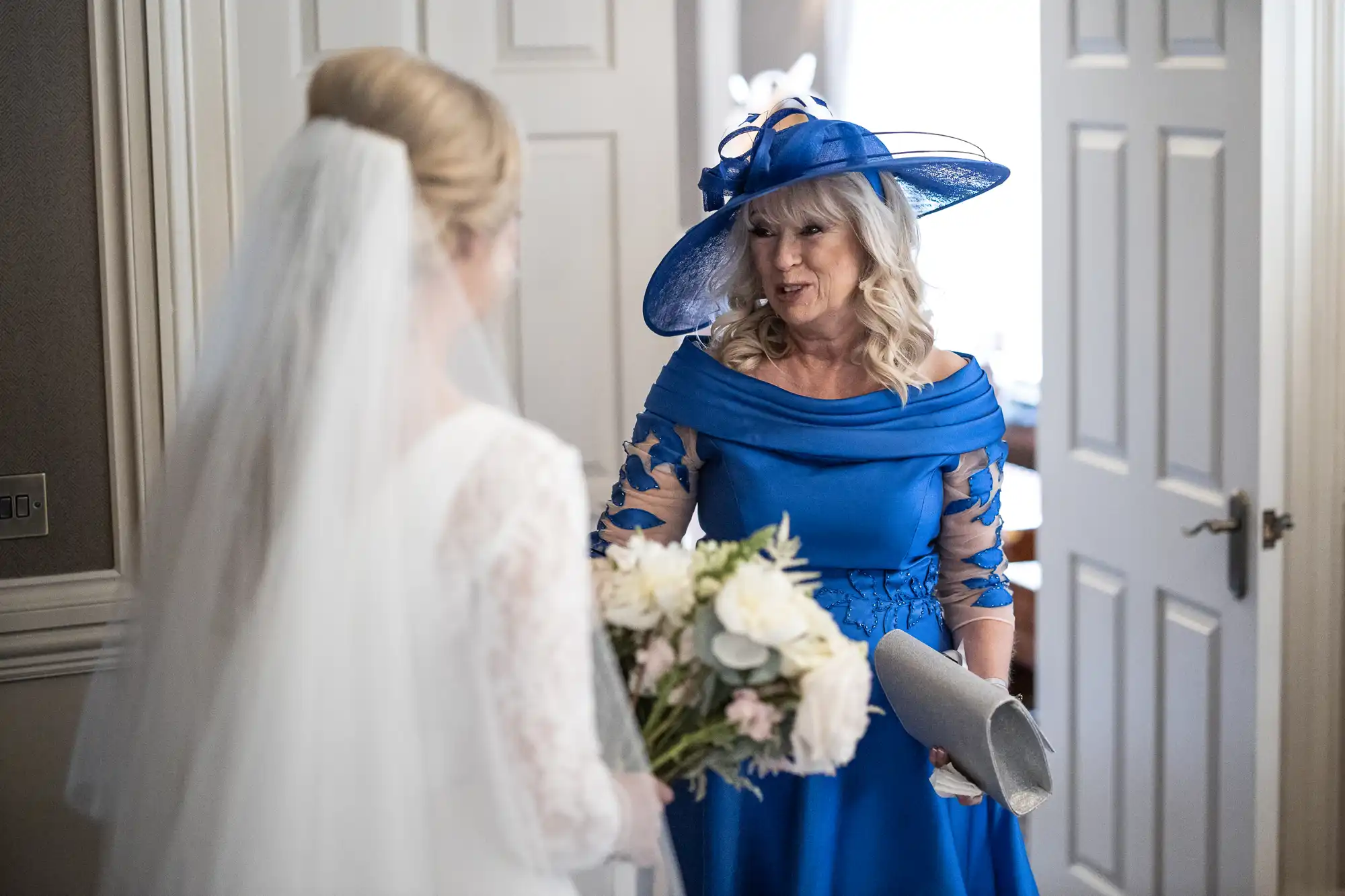 A woman in a blue dress and hat talks to a bride holding a bouquet of flowers in a room with white doors.