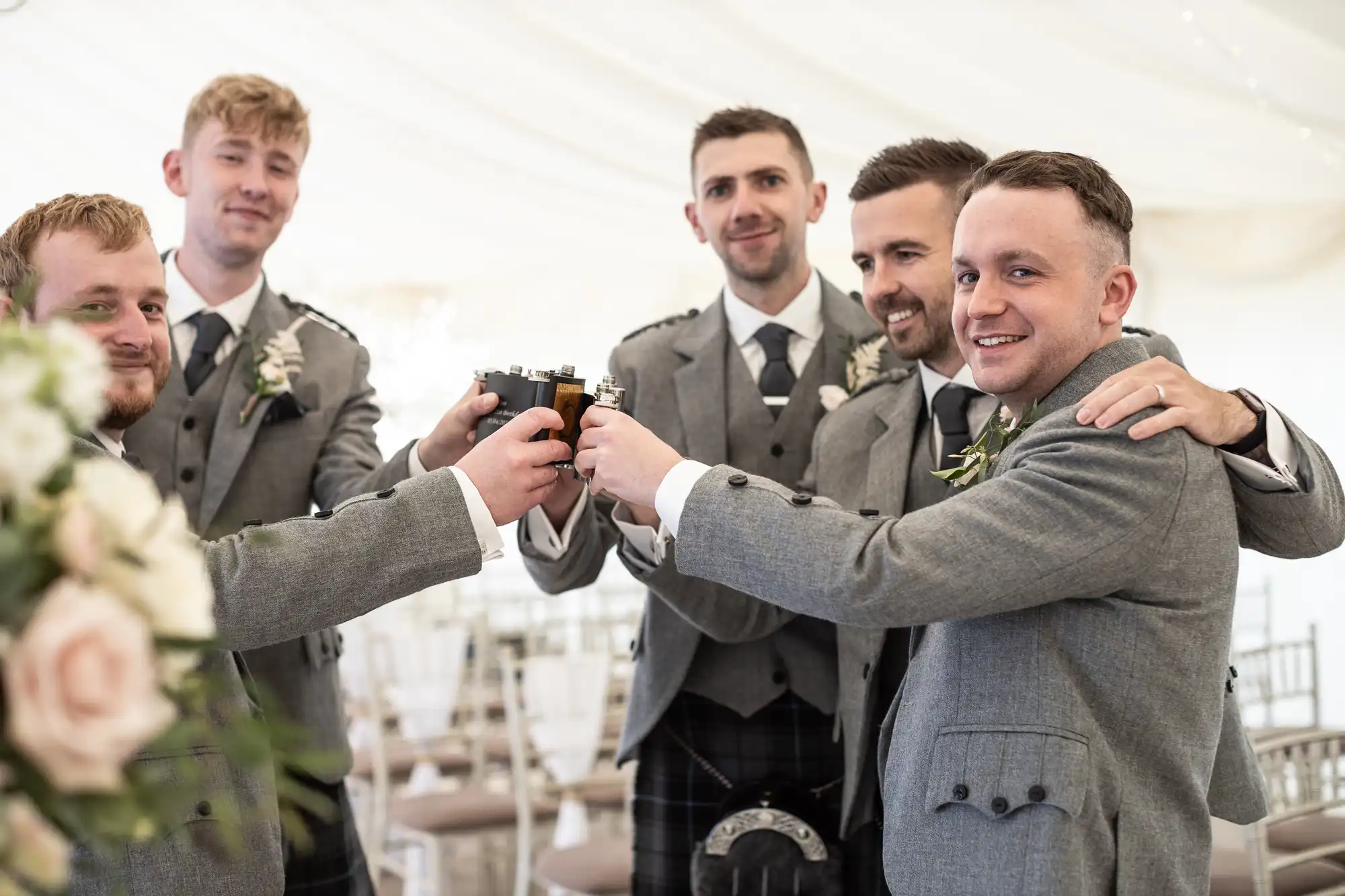 Five men in grey suits smile and raise glasses, toasting together in a decorated indoor setting.