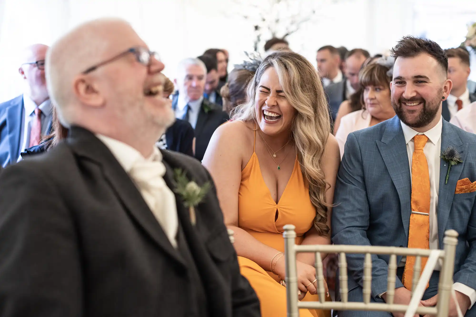 A man, woman, and another man are seated and sharing a laugh at an event, with a crowd of people in the background.