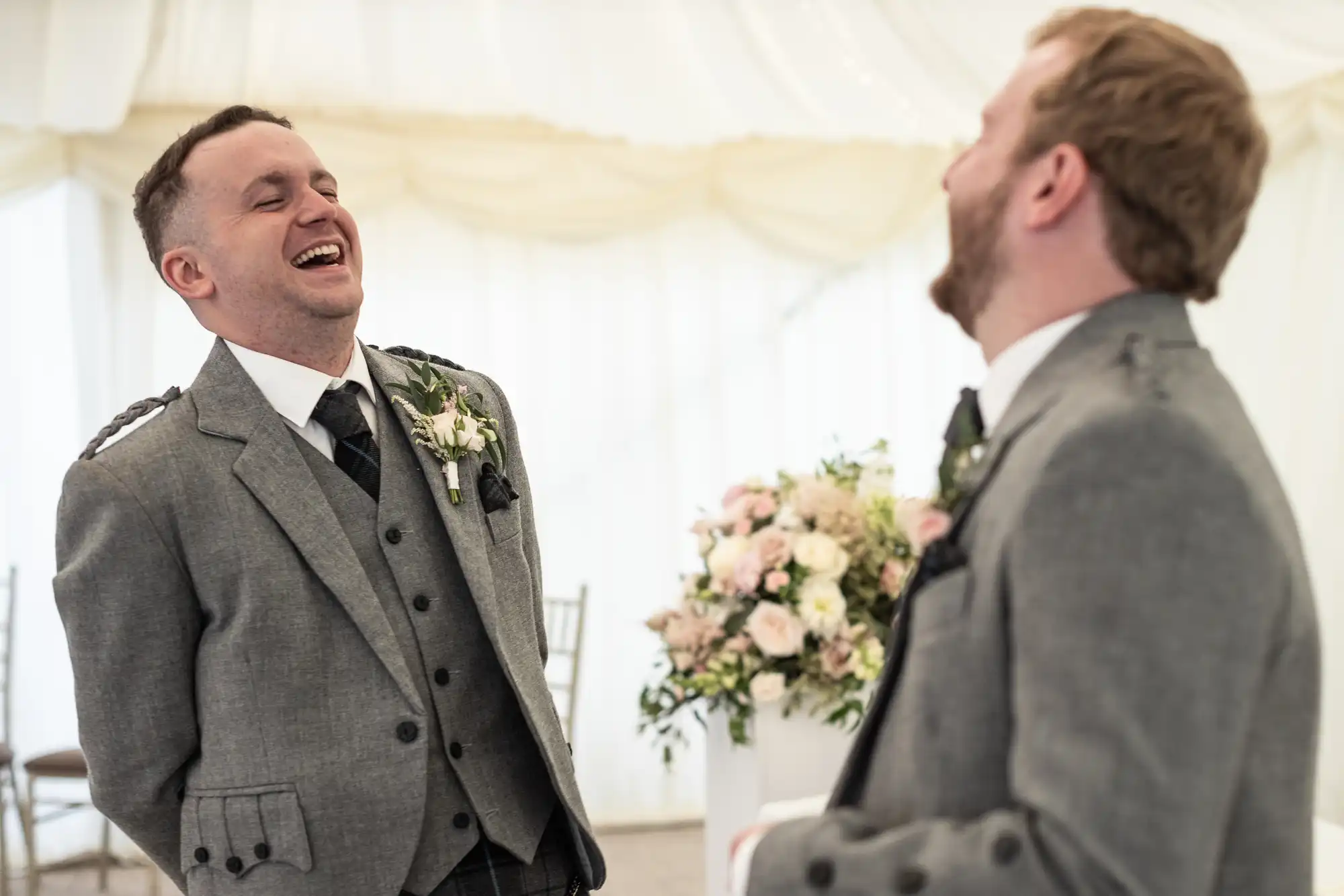 Two men in grey suits and matching ties share a hearty laugh inside a venue decorated with flowers and light-colored drapery.