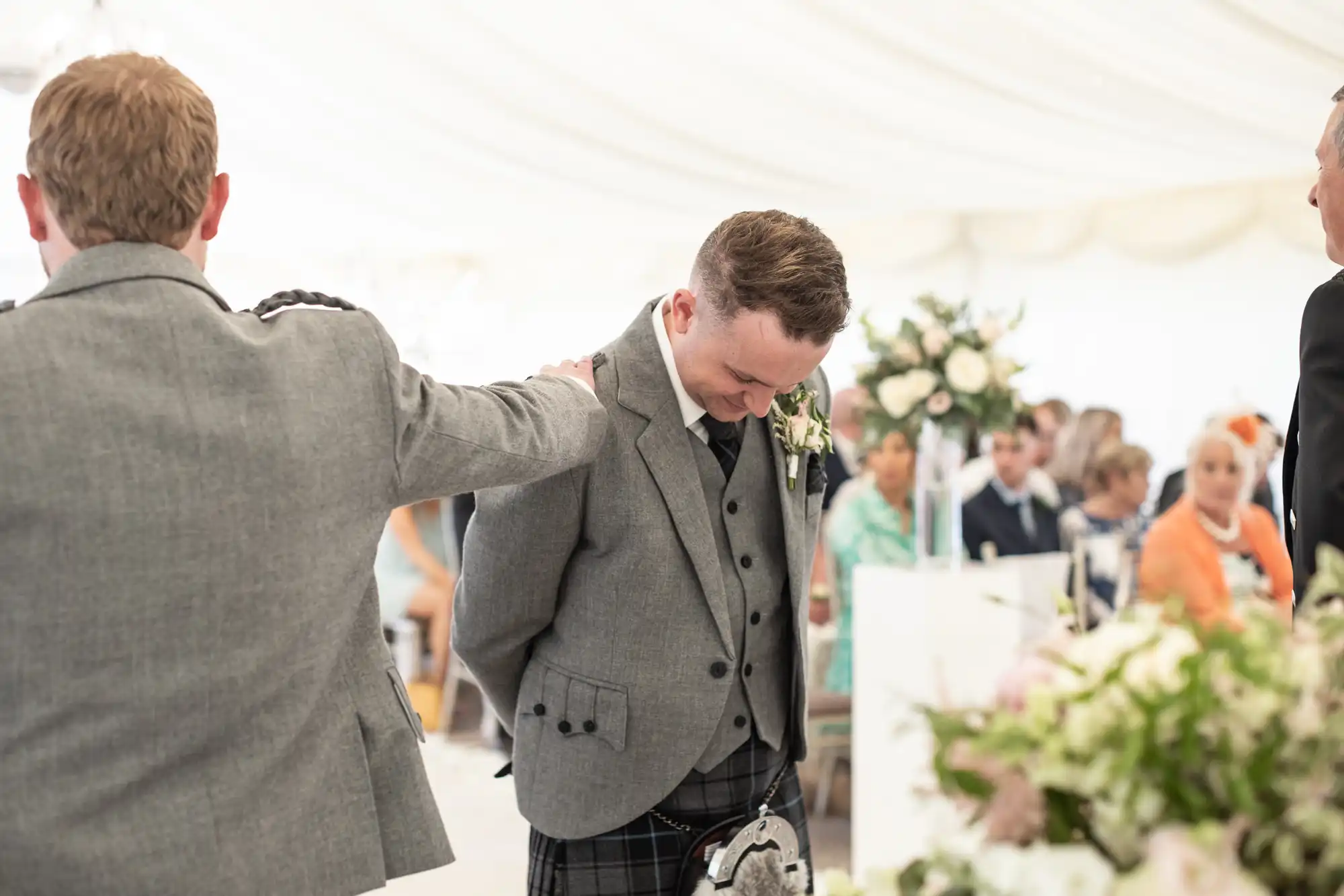 A man wearing a grey suit and plaid kilt at a wedding ceremony smiles as another man, also in a grey suit, gives him a reassuring pat on the back. Guests are seated in the background.