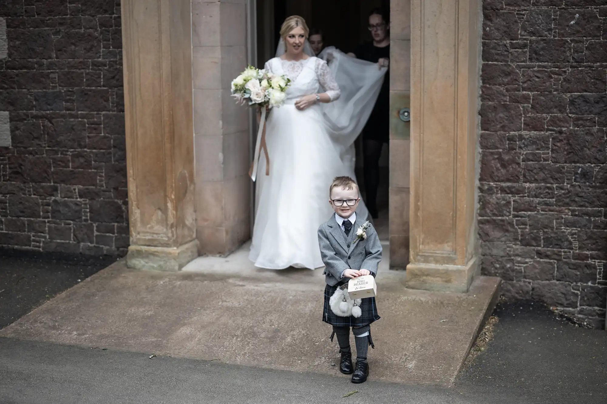 A bride holding a bouquet exits a stone building, assisted by another person. A young boy in a suit and kilt walks ahead of her, holding a small sign and a plush toy.