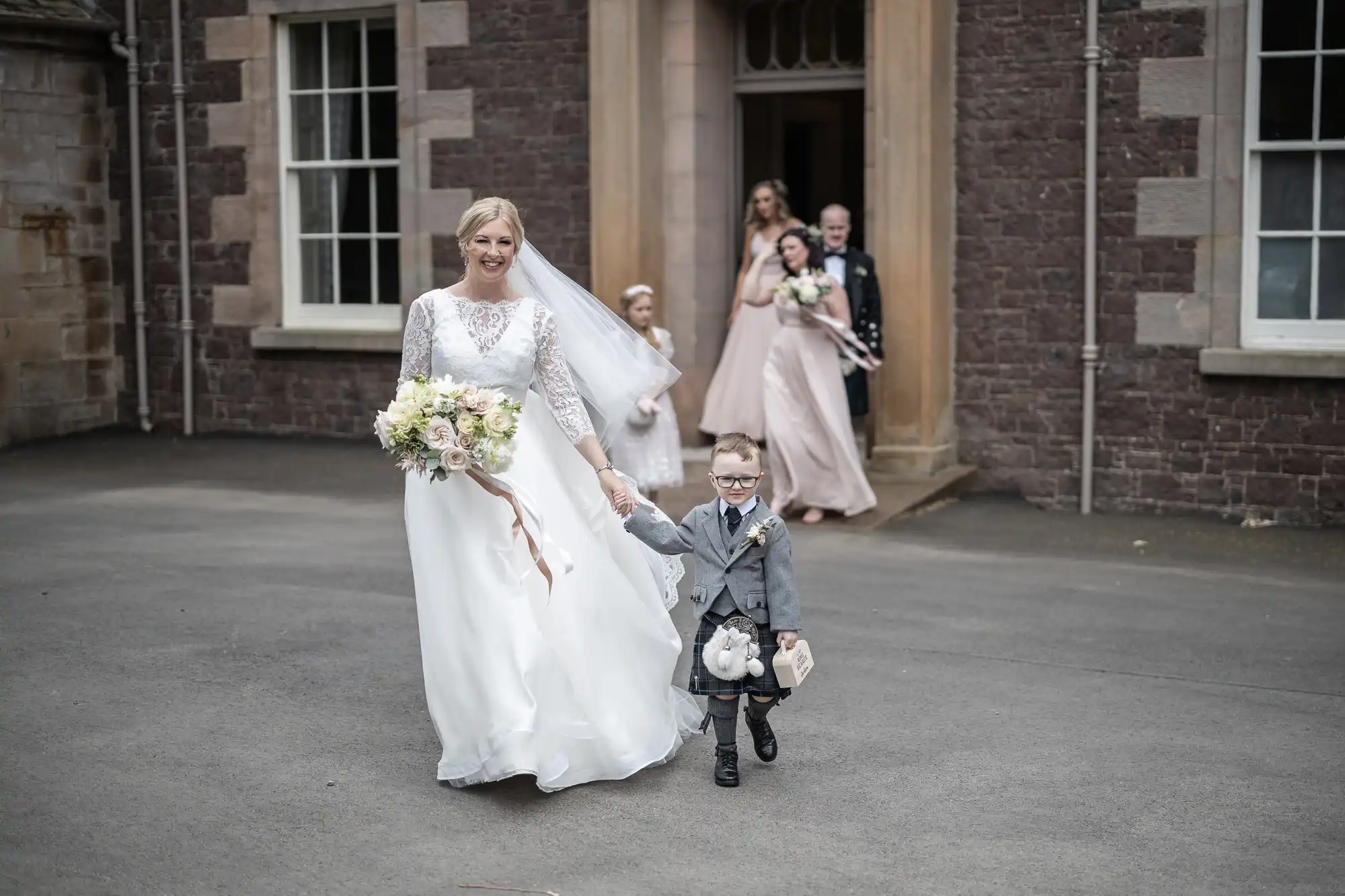 A bride holding a bouquet walks hand-in-hand with a young boy dressed in a gray suit while other wedding party members follow behind outside a stone building.