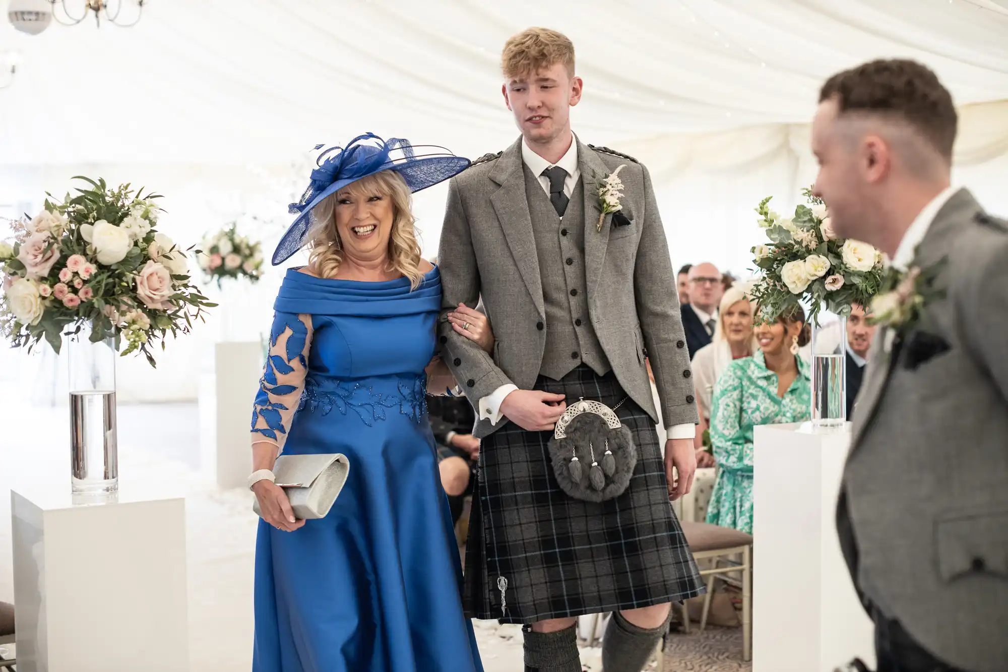 Woman in a blue dress and hat escorting a man in a gray jacket and kilt down a wedding aisle, with guests seated on either side.