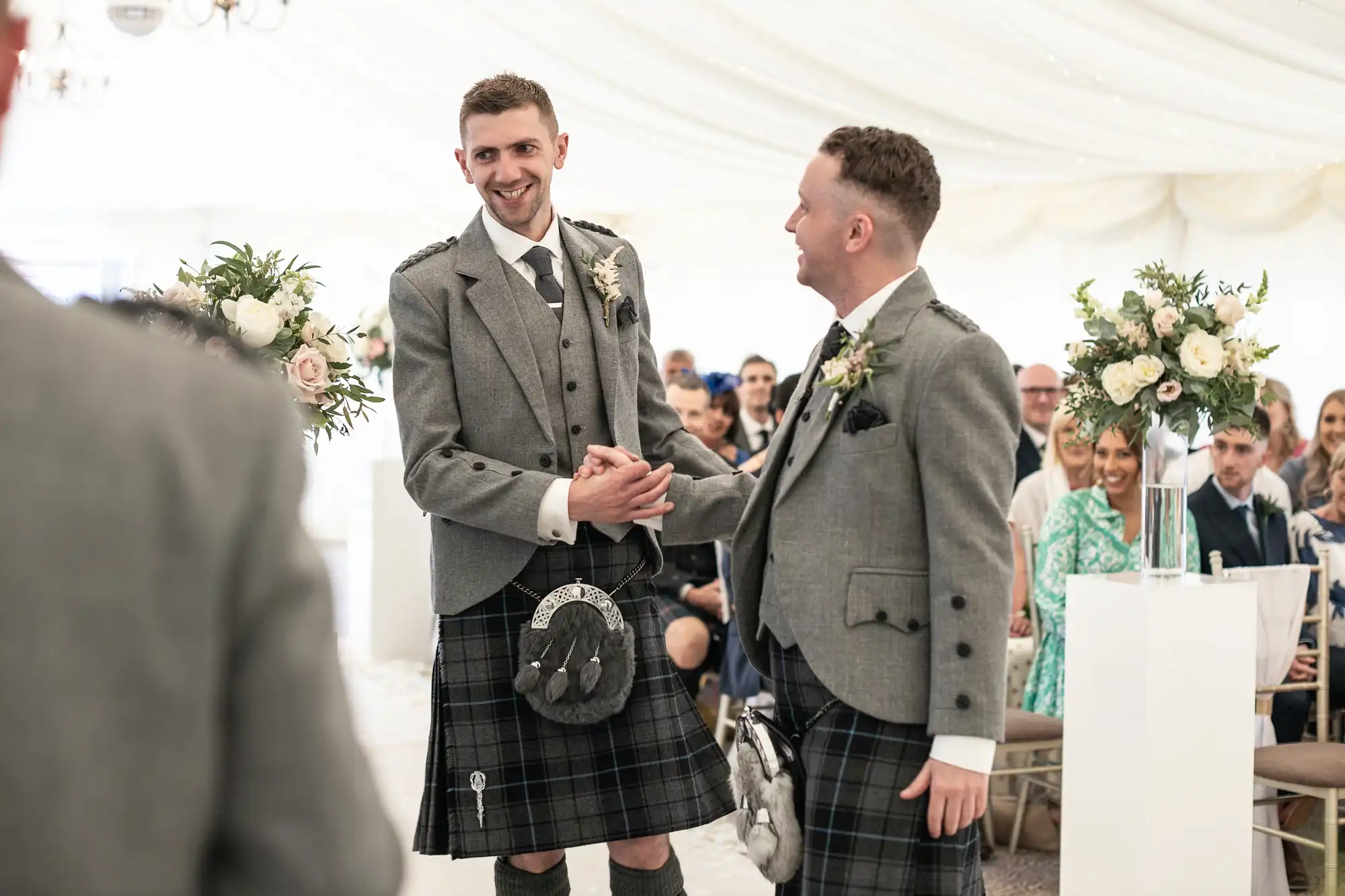 Two men in grey jackets and kilts stand together smiling, surrounded by wedding guests in a decorated venue with floral arrangements.