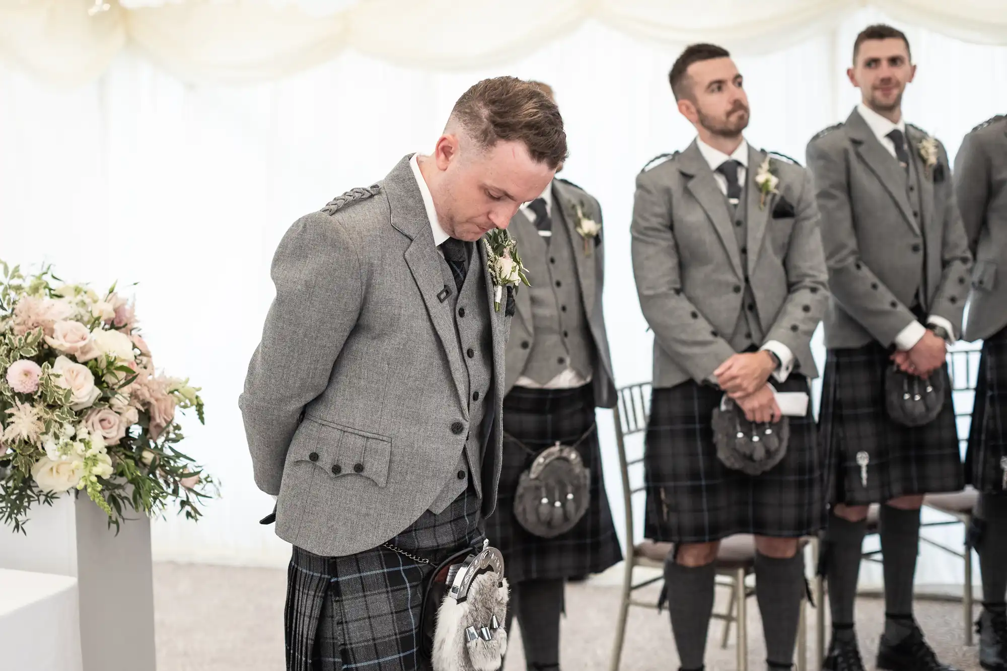 A man in traditional Scottish attire stands looking down with hands behind his back. Three other men in similar attire stand in the background. A floral arrangement is partially visible on the left.