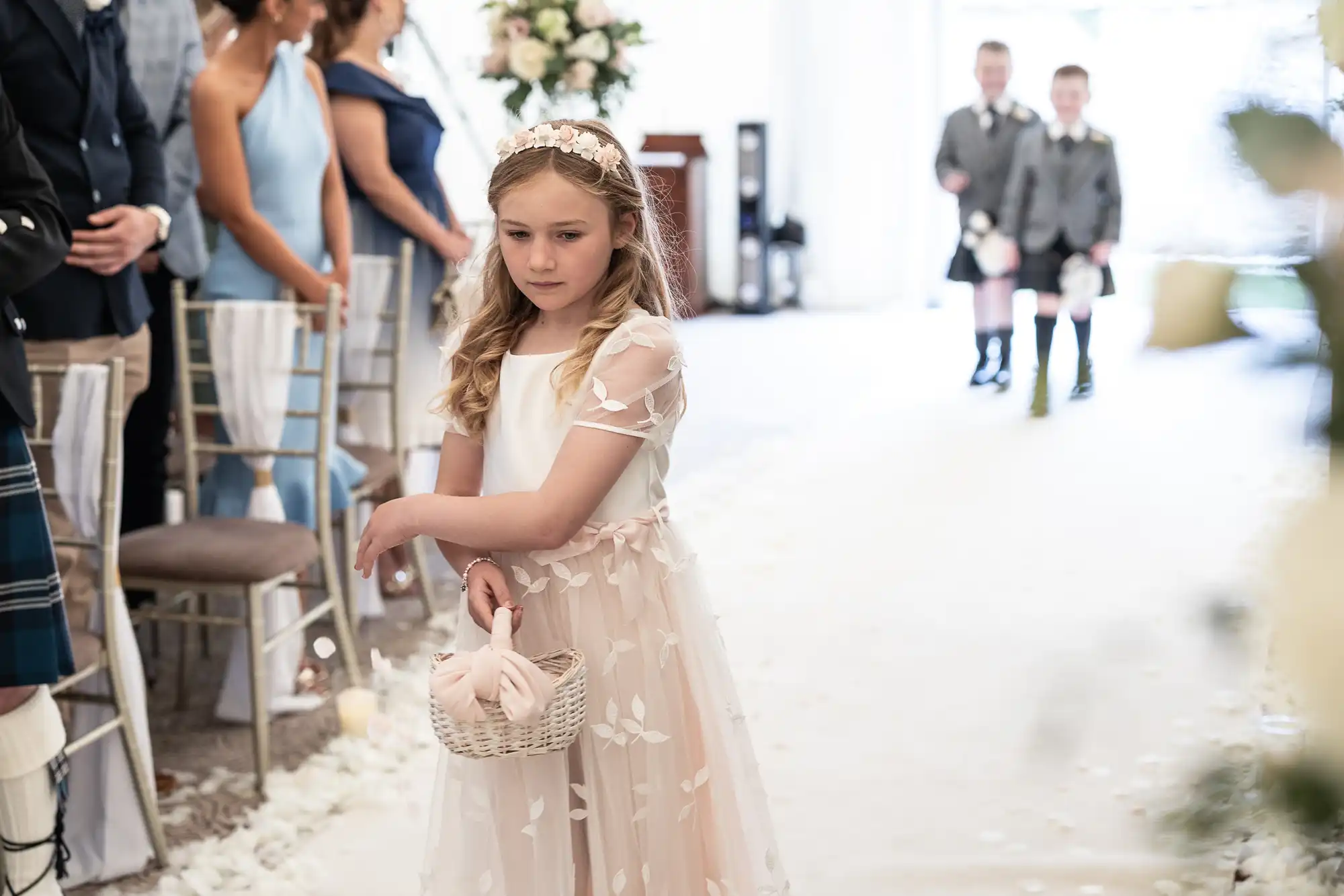 A young girl dressed in a white dress walks down an aisle, sprinkling petals from a basket. She is part of a ceremony with guests seated on either side and two young boys in the background.