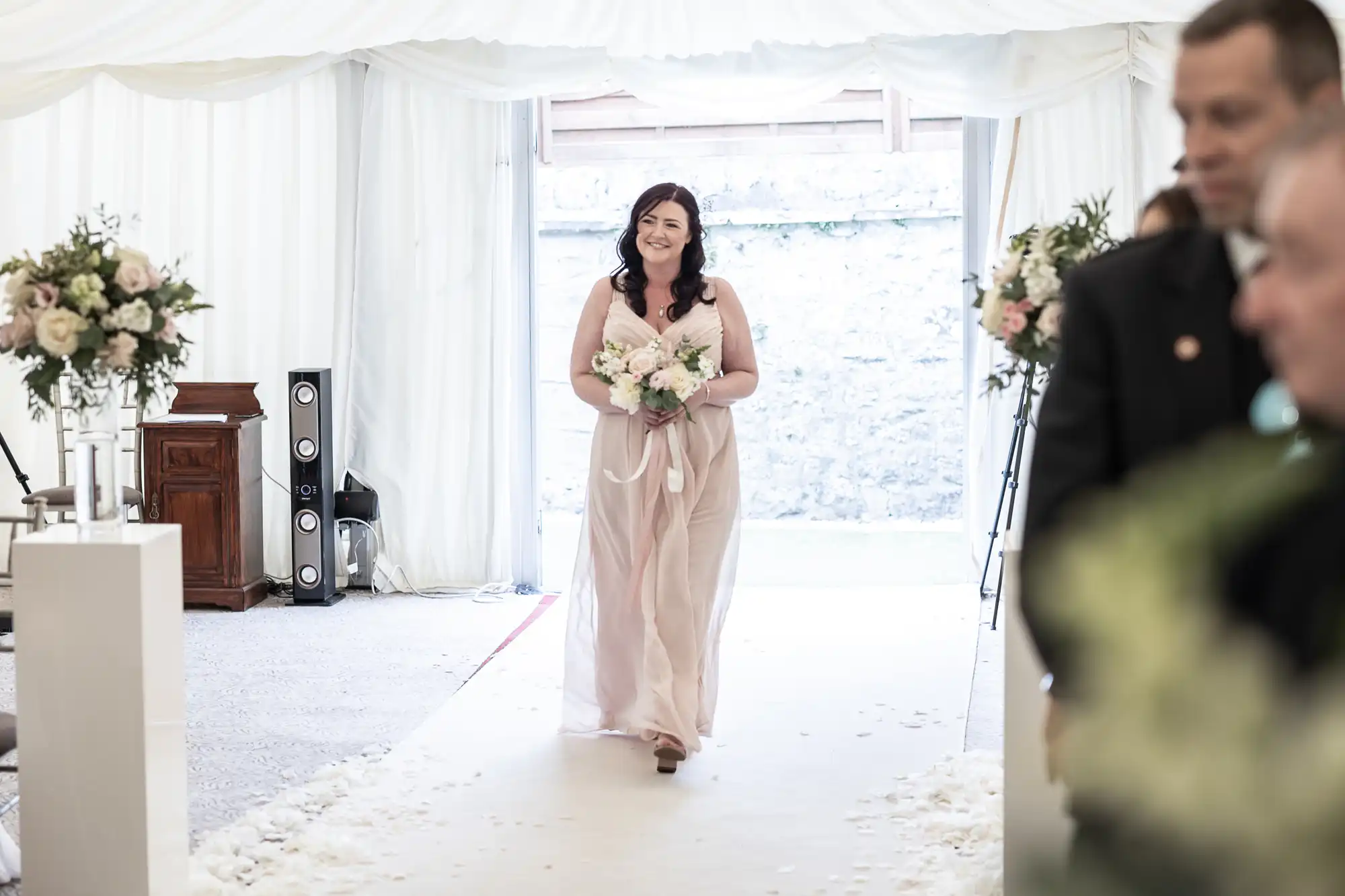 A woman in a light pink dress walks down an aisle, holding a bouquet of flowers, in a decorated event tent.